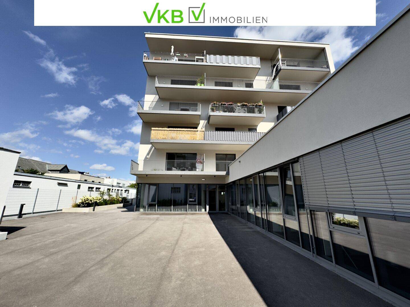 Ansicht eines modernen Gebäudes mit großen Glasflächen im Erdgeschoss und weitläufigem Vorplatz.