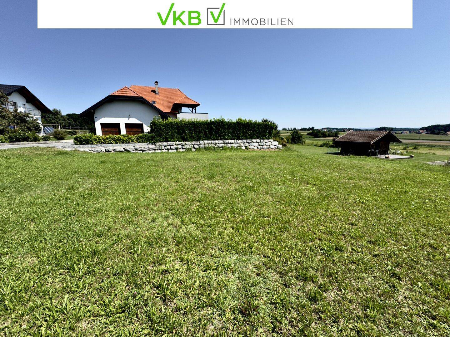 Offene Rasenfläche mit Blick auf Wohnhäuser und ein kleines Gartenhaus in der Ferne bei strahlendem Wetter.
