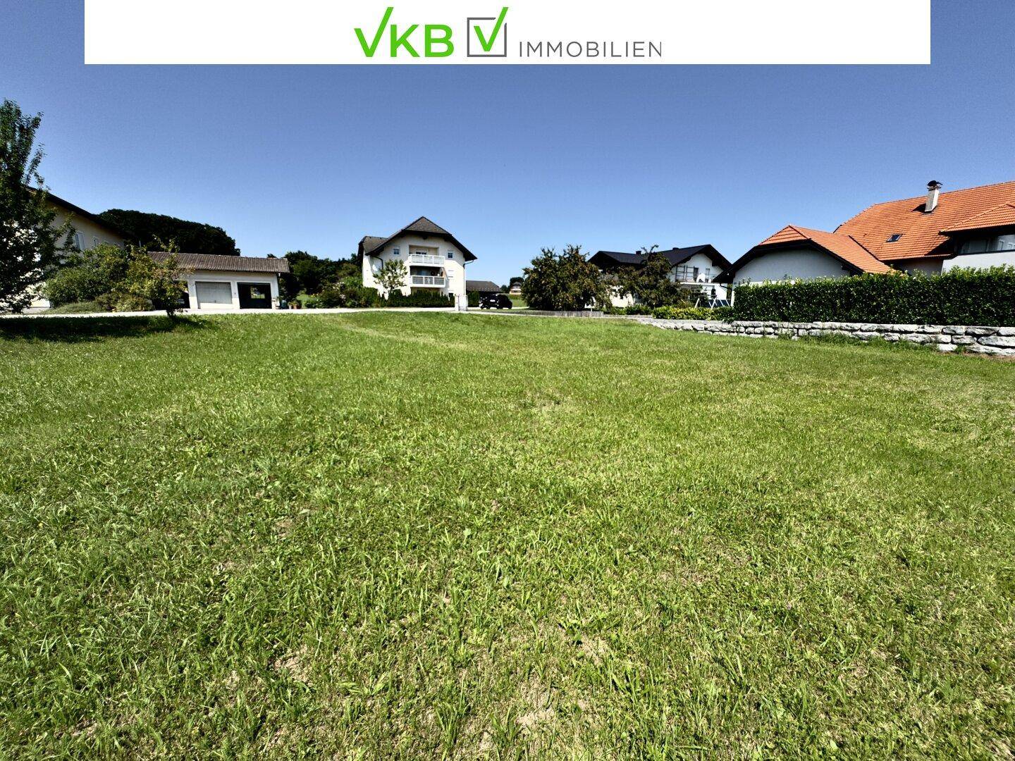 Weitläufige Grünfläche mit mehreren Wohnhäusern und einer Hecke im Hintergrund unter blauem Himmel.