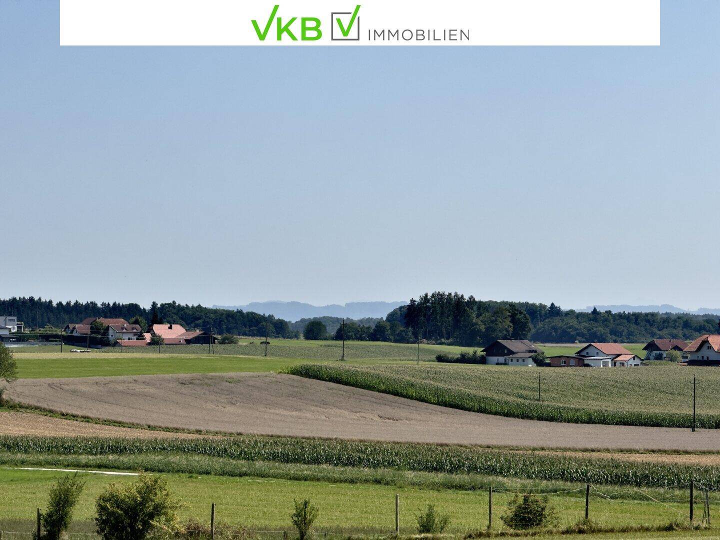 Panoramablick über weite Felder, ländliche Bebauung und eine hügelige Landschaft am Horizont an einem Sommertag.