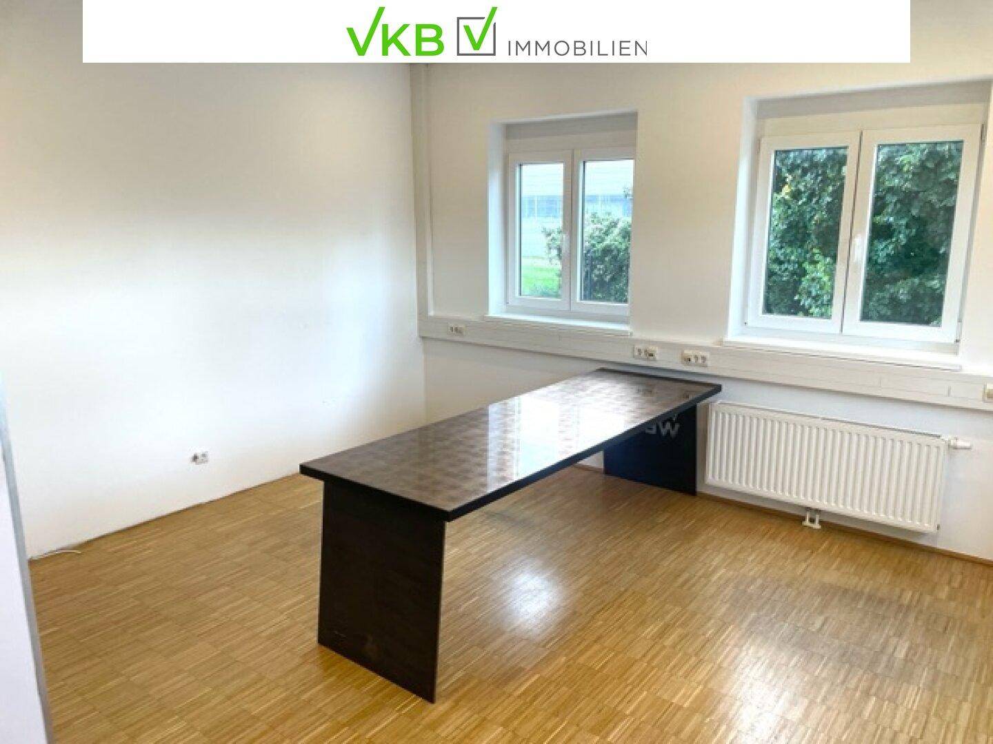 Leeres Büro mit einem langen dunklen Tisch, zwei Fenstern und einem Heizkörper, bereit zur Einrichtung.