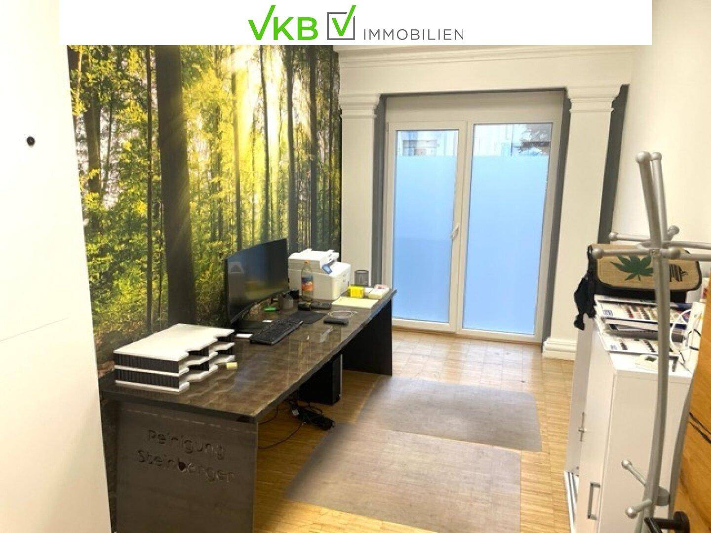 Kleines Büro mit Schreibtisch, Computer und Drucker, beleuchtet durch eine große Fensterfront mit Waldmotiv.