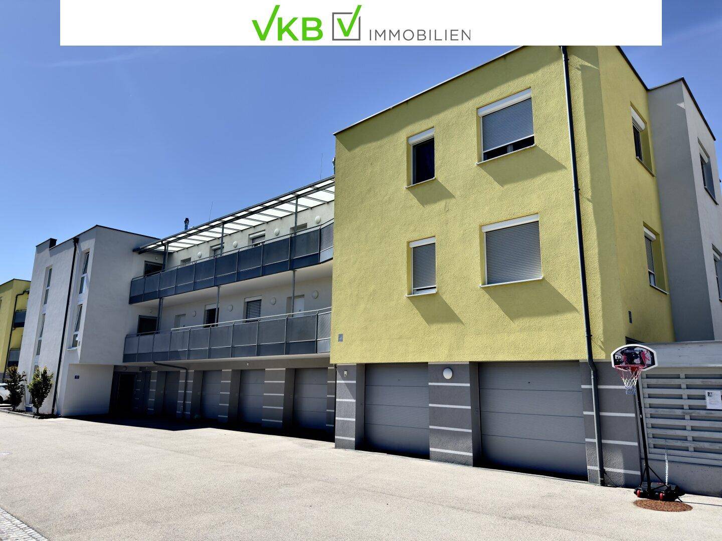Modernes Wohngebäude mit gelber Fassade, Balkonen, Garagen und einem Basketballkorb im Vordergrund.