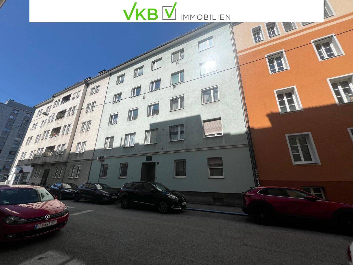 Mehrstöckiges Wohngebäude an einer Straße mit geparkten Autos unter blauem Himmel.