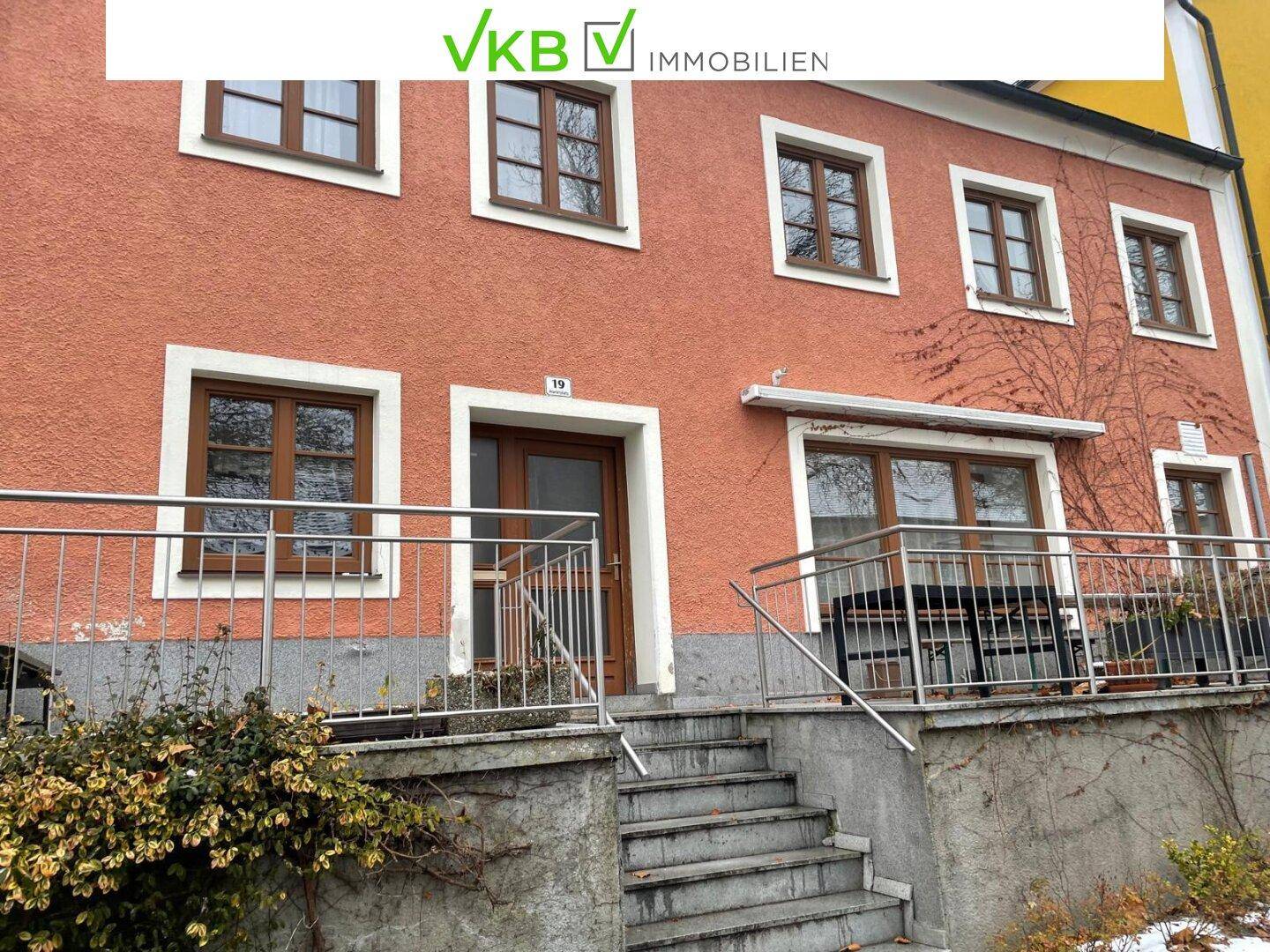 Fassade eines Wohnhauses mit orangeroter Farbe, Holzfenstern und einem Eingangsbereich mit Metallgeländer.