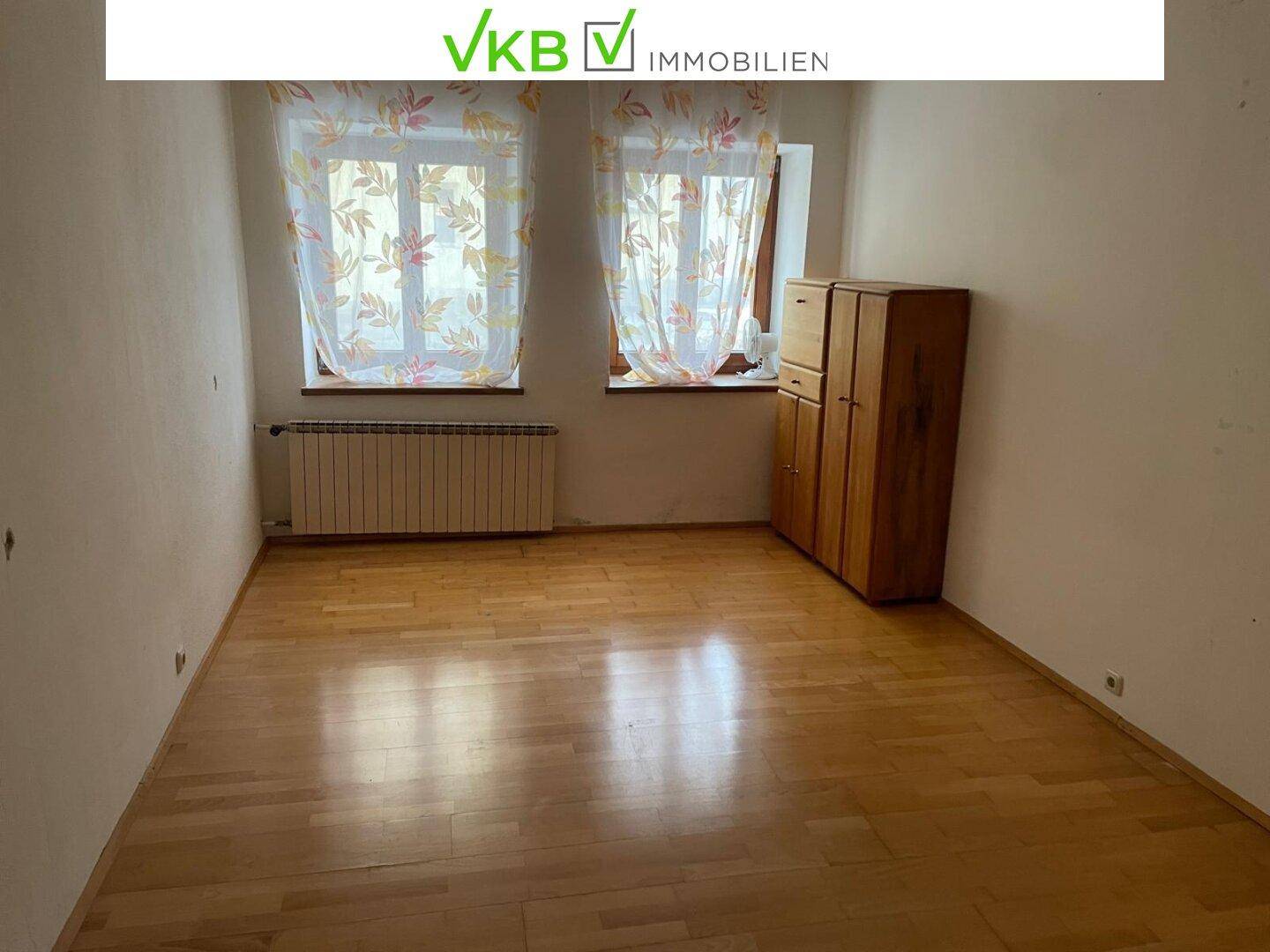 Helles Schlafzimmer mit Holzboden, zwei Fenstern und einem Heizkörper, einfach eingerichtet.