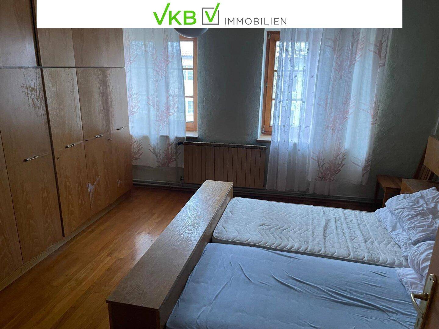 Schlafzimmer mit Holzboden, zwei Fenstern und einem großen Kleiderschrank, einfach eingerichtet.