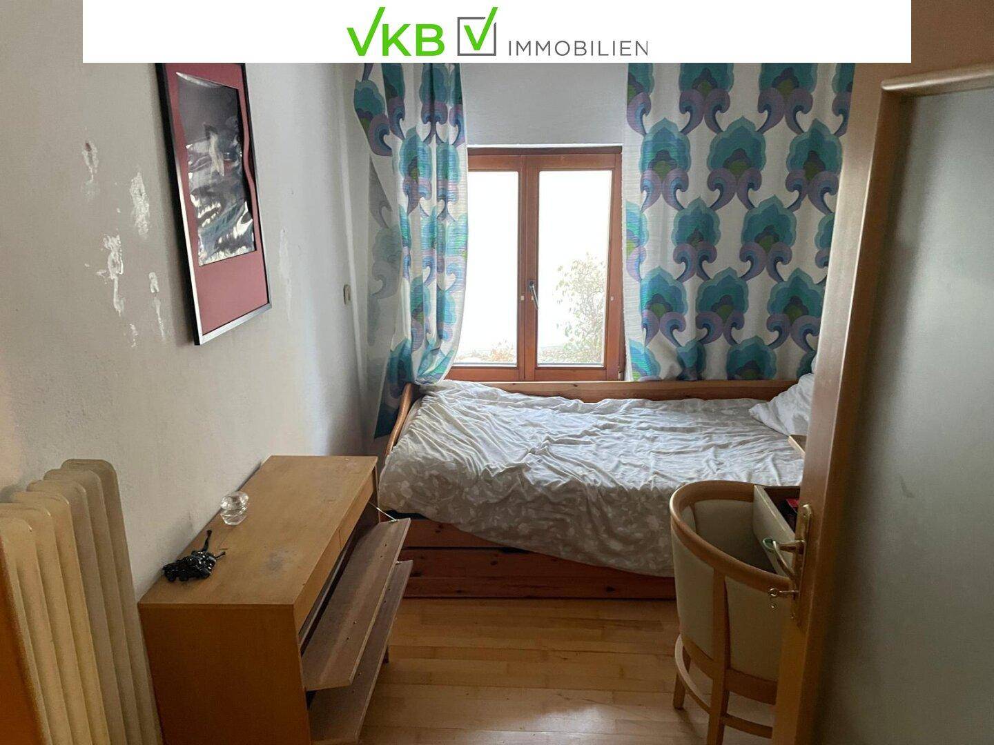 Kleines Schlafzimmer mit Holzboden, einem Bett und einem Fenster mit gemusterten Vorhängen.