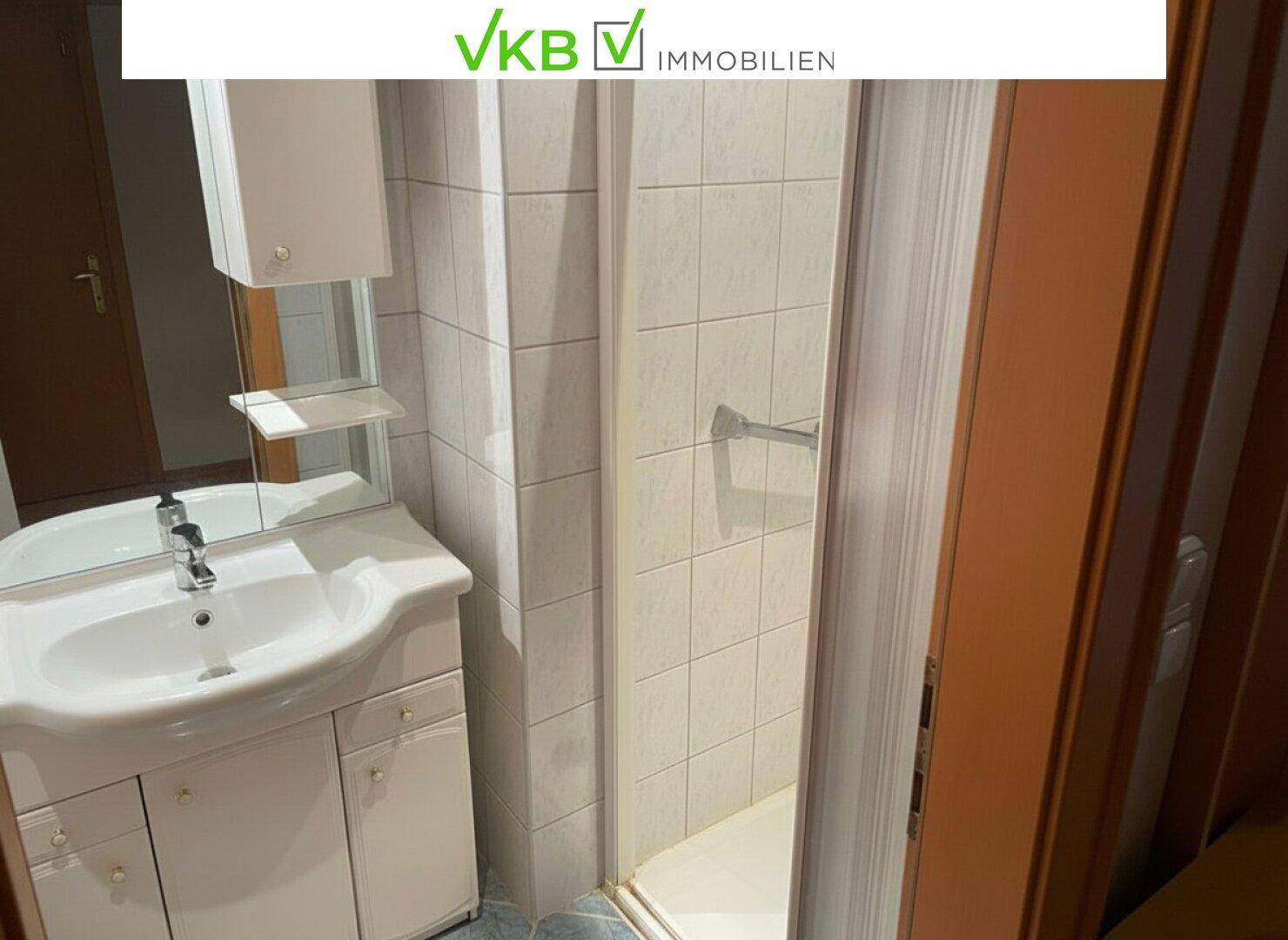 Badezimmer mit Duschkabine, Waschbecken und Spiegel, ausgestattet mit weißen Fliesen und einfacher Einrichtung.