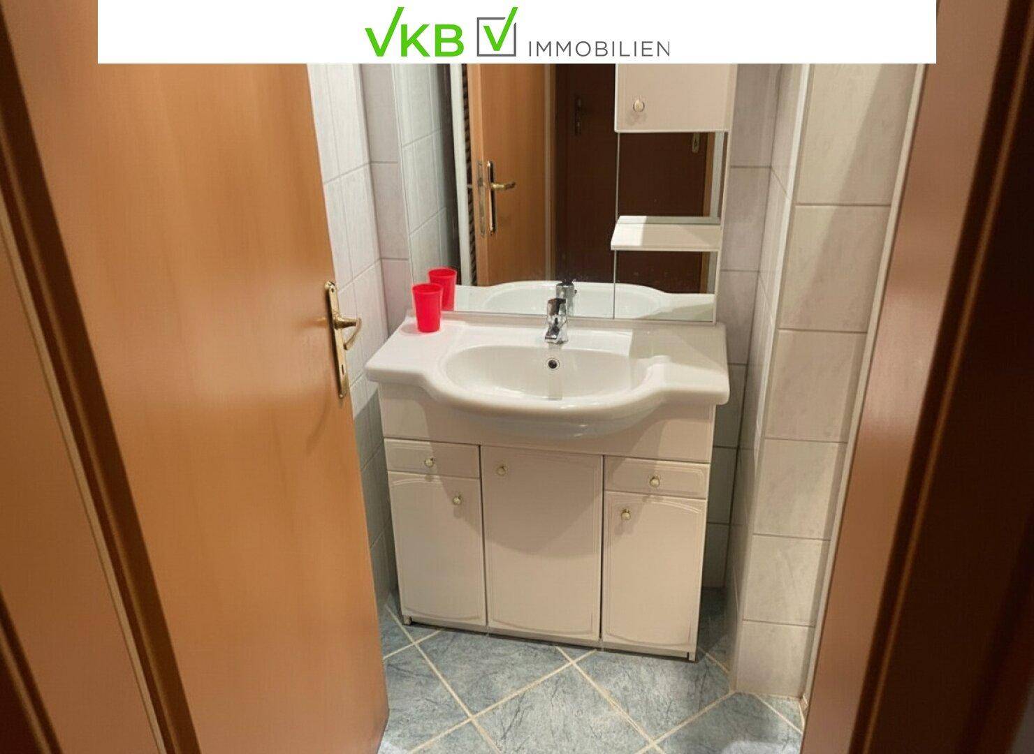 Badezimmer mit Waschbecken, Unterschrank und Spiegel, umgeben von weißen Fliesen und einer Holztür.
