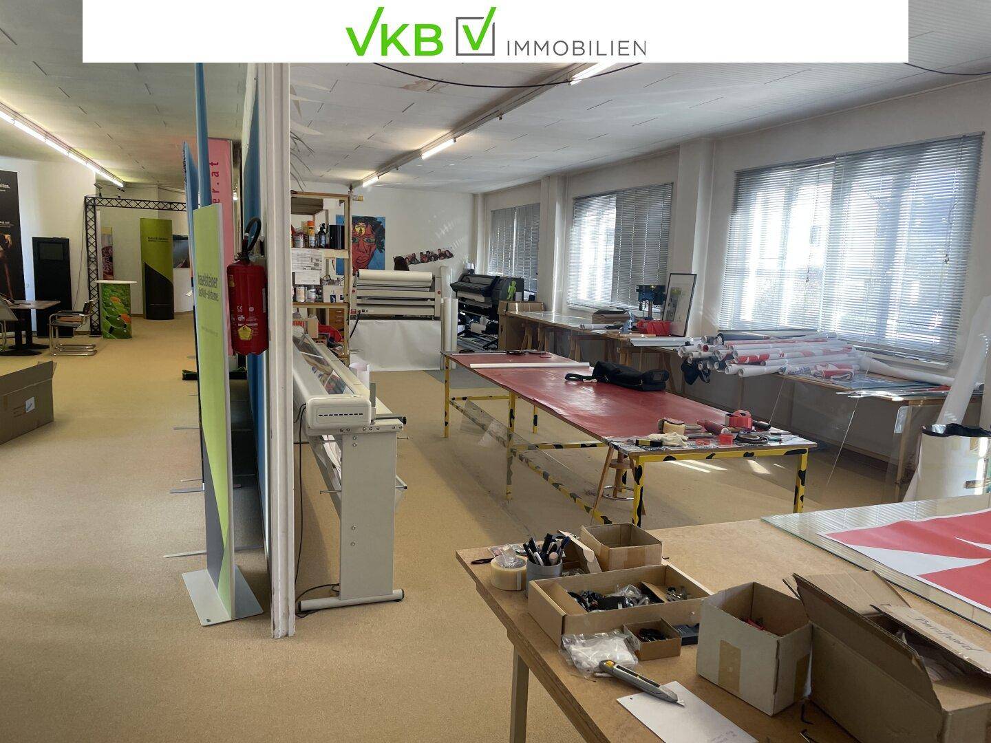 Heller Arbeitsbereich mit großen Fenstern, Teppichboden und verschiedenen Tischen und Geräten.