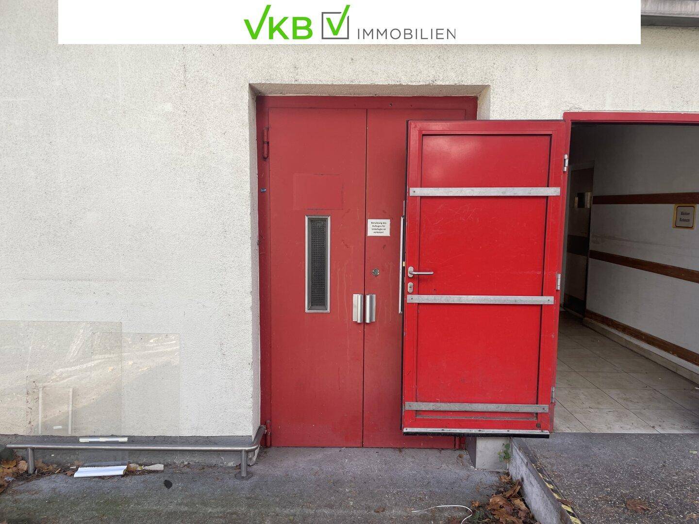 Rote Doppeltür als Zugang zum Gebäude, eine davon offen, mit einfacher Betonumgebung.