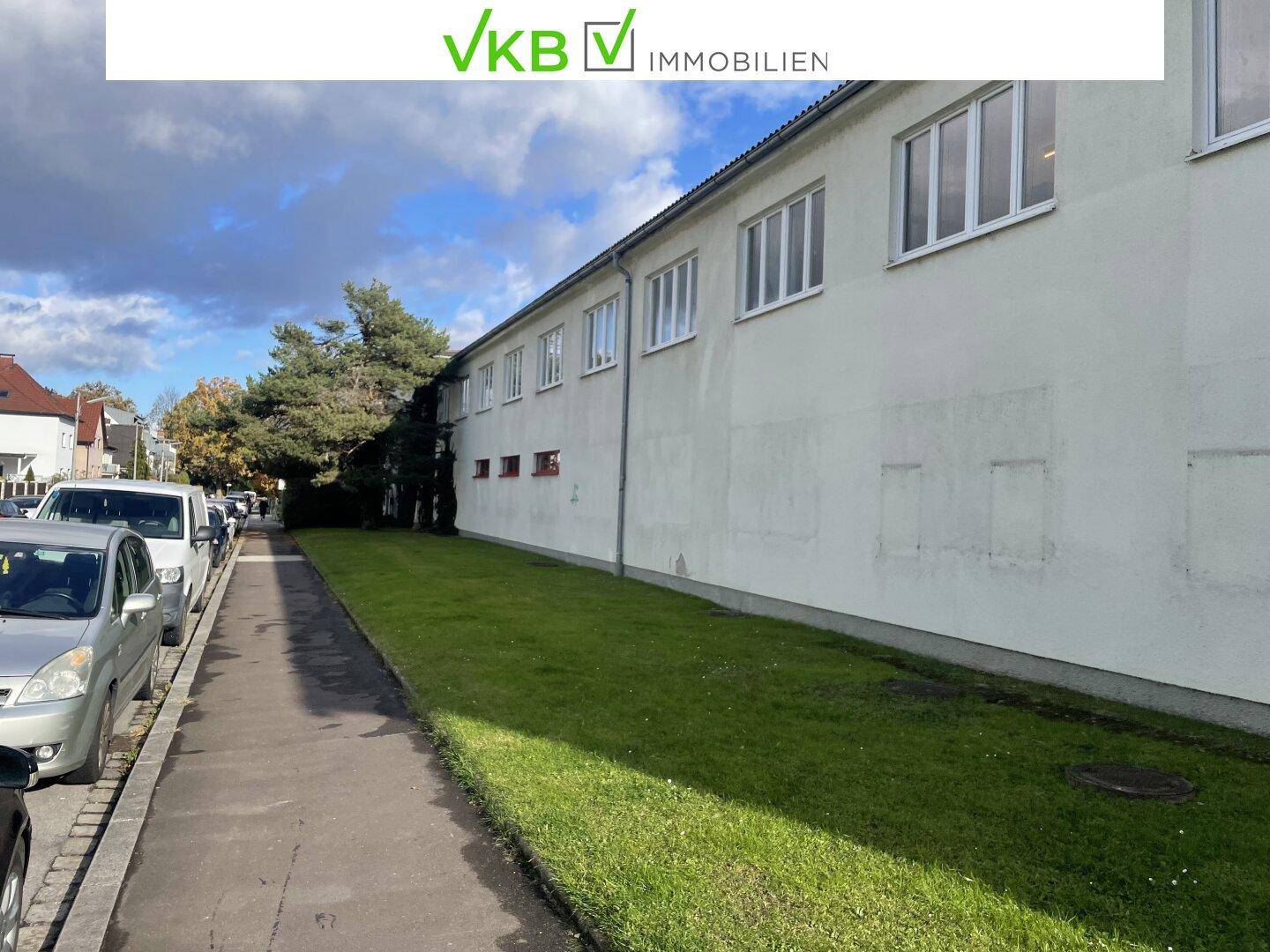 Seitenansicht des Gebäudes mit einer Grünfläche entlang der Fassade und parkenden Autos auf der Straße.