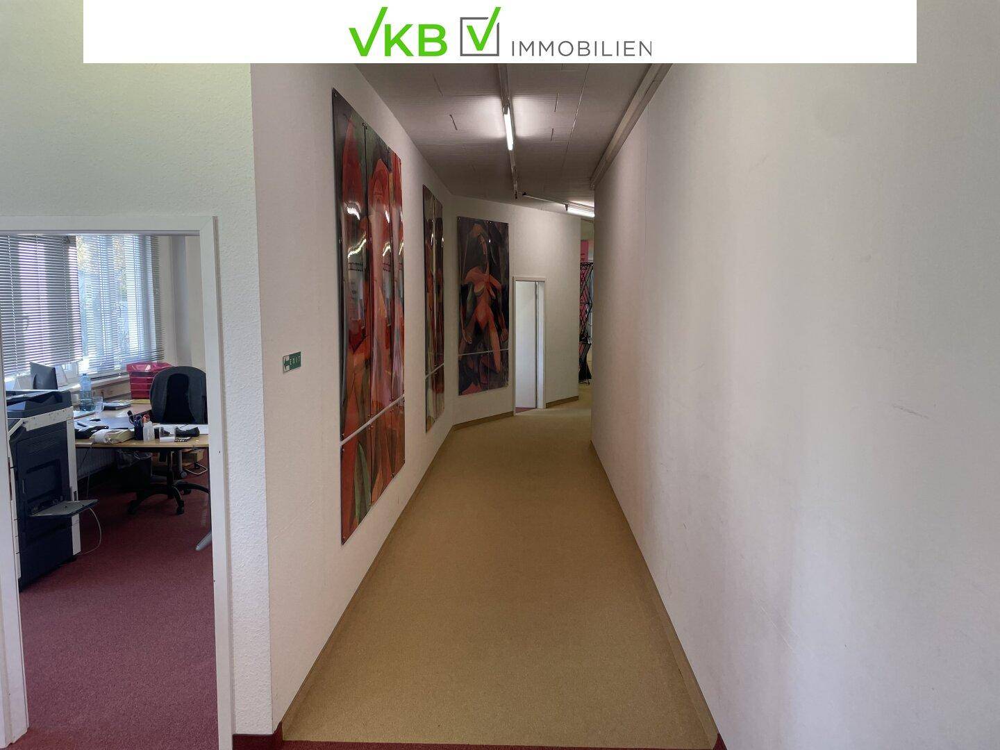 Langer Flur mit Teppichboden und Kunstwerken an den Wänden, der zu Büroräumen führt.