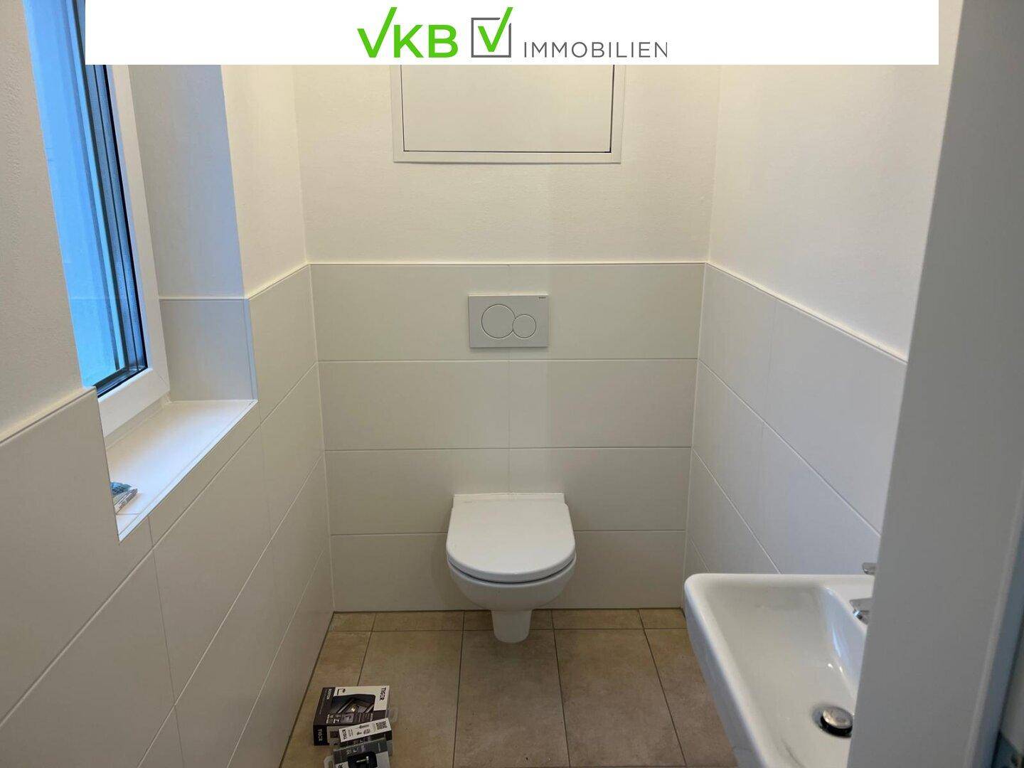 Modernes Gäste-WC mit weißen Fliesen, wandhängendem WC, Waschbecken und Fenster.