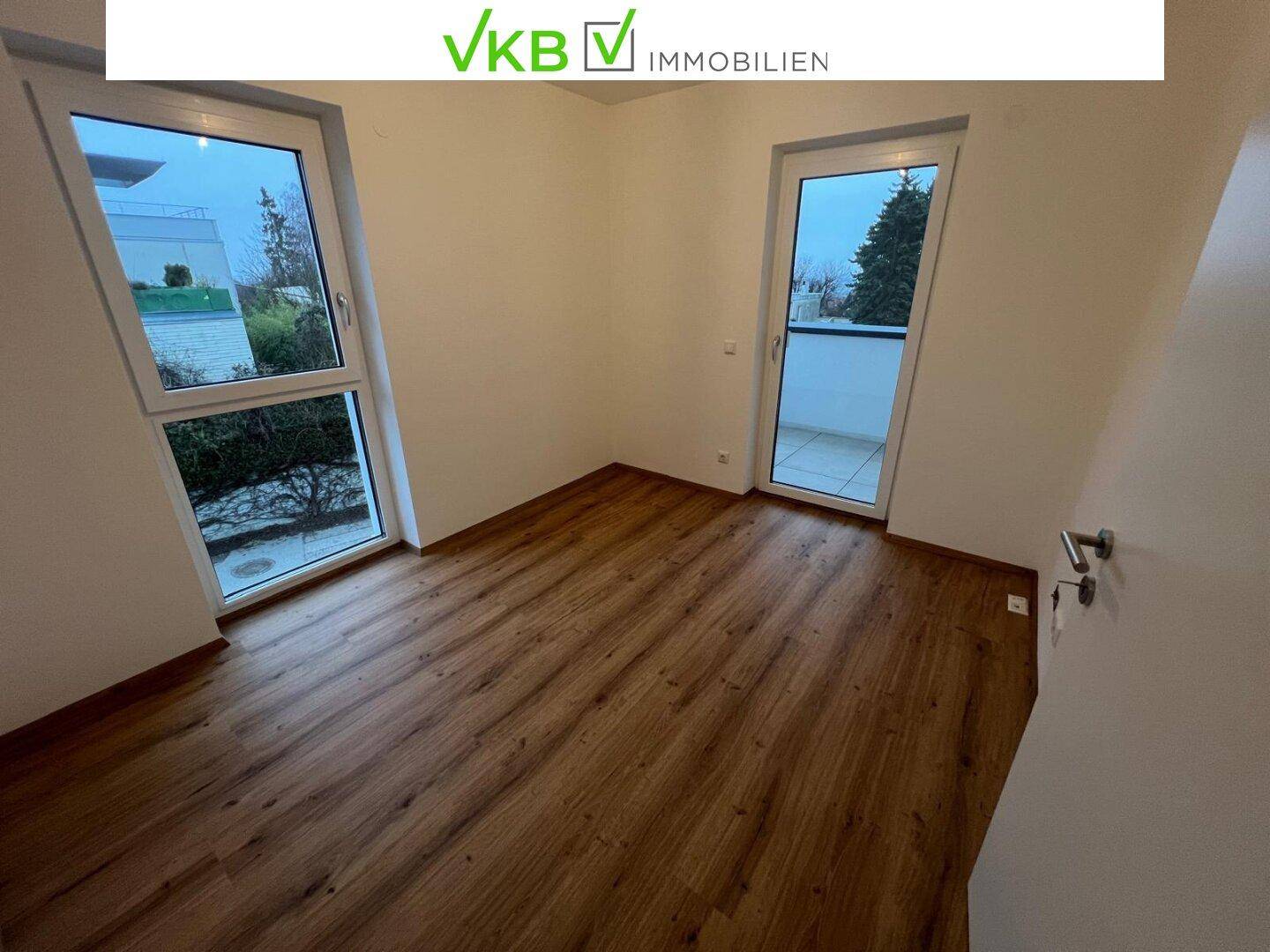 Helles Zimmer mit Holzboden, zwei Fenstern und Zugang zu einem kleinen Balkon.