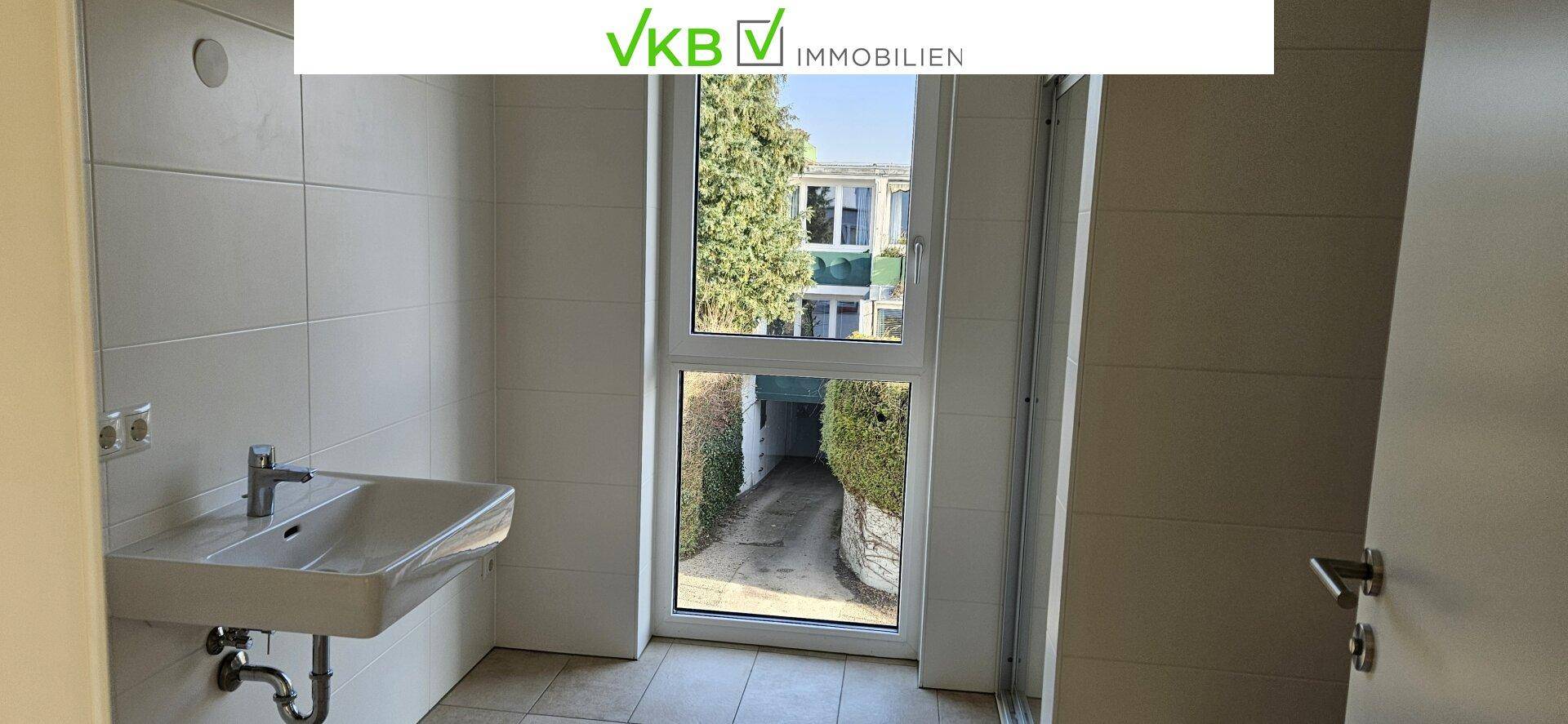 Helles Badezimmer mit weißen Fliesen, Waschbecken und Fenster mit Blick auf die Einfahrt.