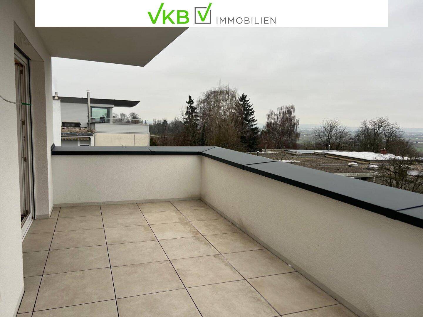 Geräumige, geflieste Terrasse mit weißer Brüstung und Blick auf Bäume und umliegende Gebäude.
