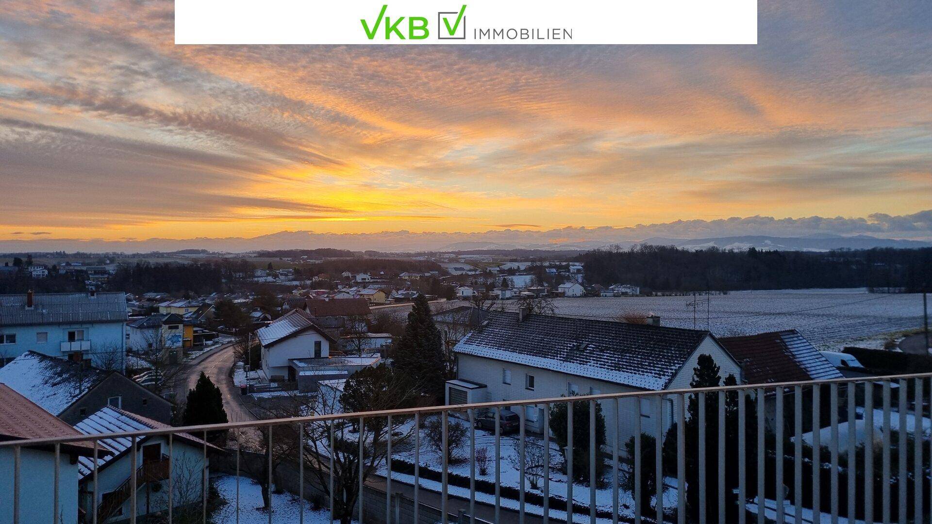 Blick von einem Balkon über eine verschneite Landschaft mit Häusern und einem farbenprächtigen Sonnenuntergang.