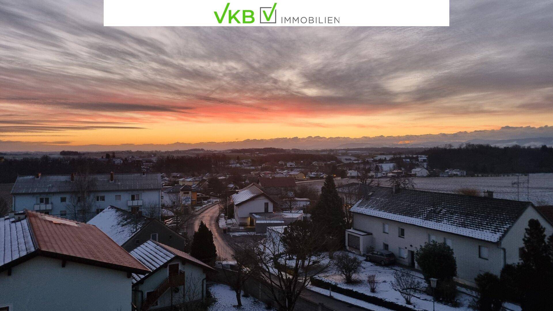 Panoramablick über eine verschneite Landschaft mit Häusern bei Sonnenuntergang und dramatischem Himmel.