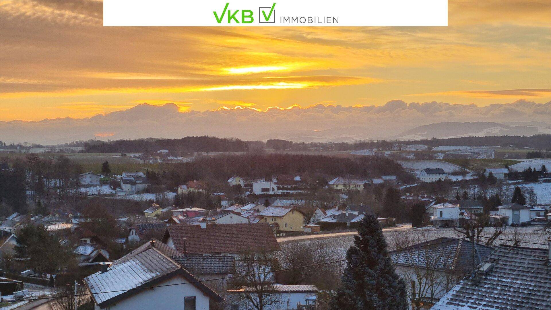 Panoramablick über eine verschneite Landschaft und Siedlung mit Bergen im Hintergrund bei Sonnenaufgang.