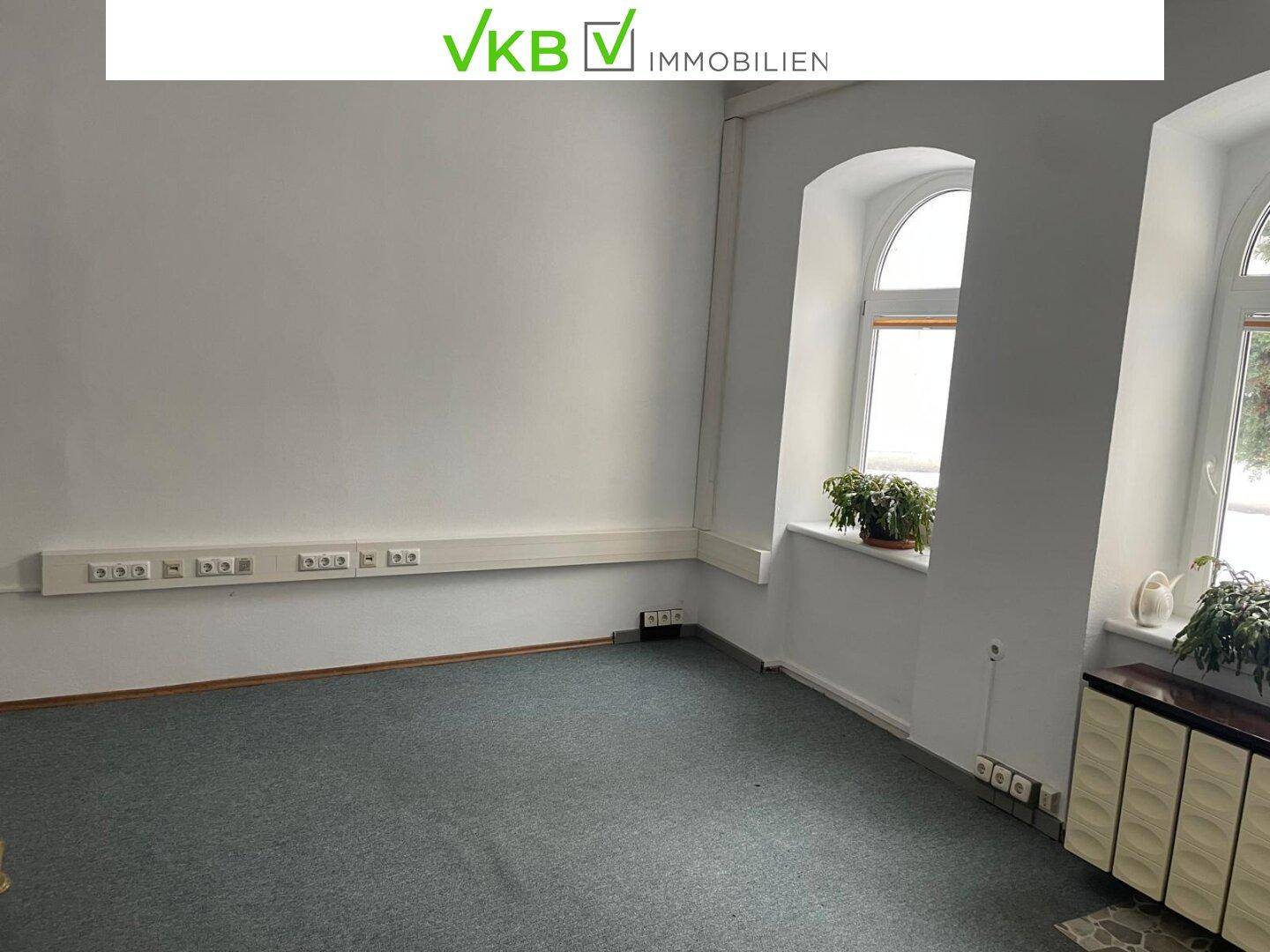 Leerer Raum mit Teppichboden, großen Fenstern und vielen Steckdosenleisten an der Wand.