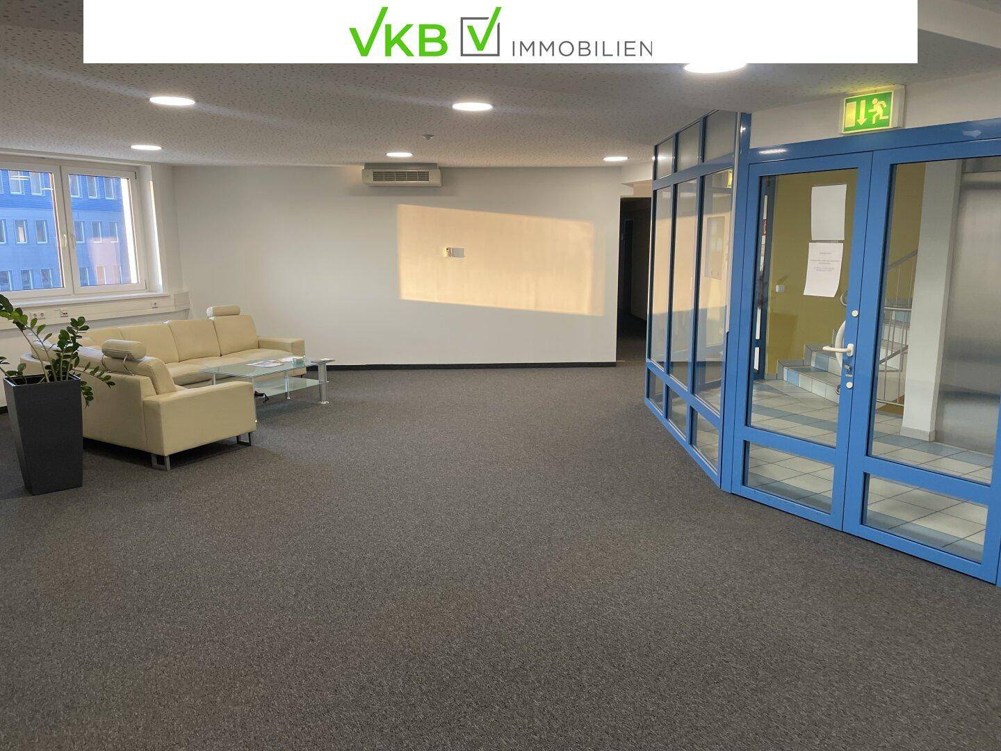Geräumiger Empfangsbereich mit beigem Sofa, Teppichboden und großen Fenstern, die viel Tageslicht hereinlassen.