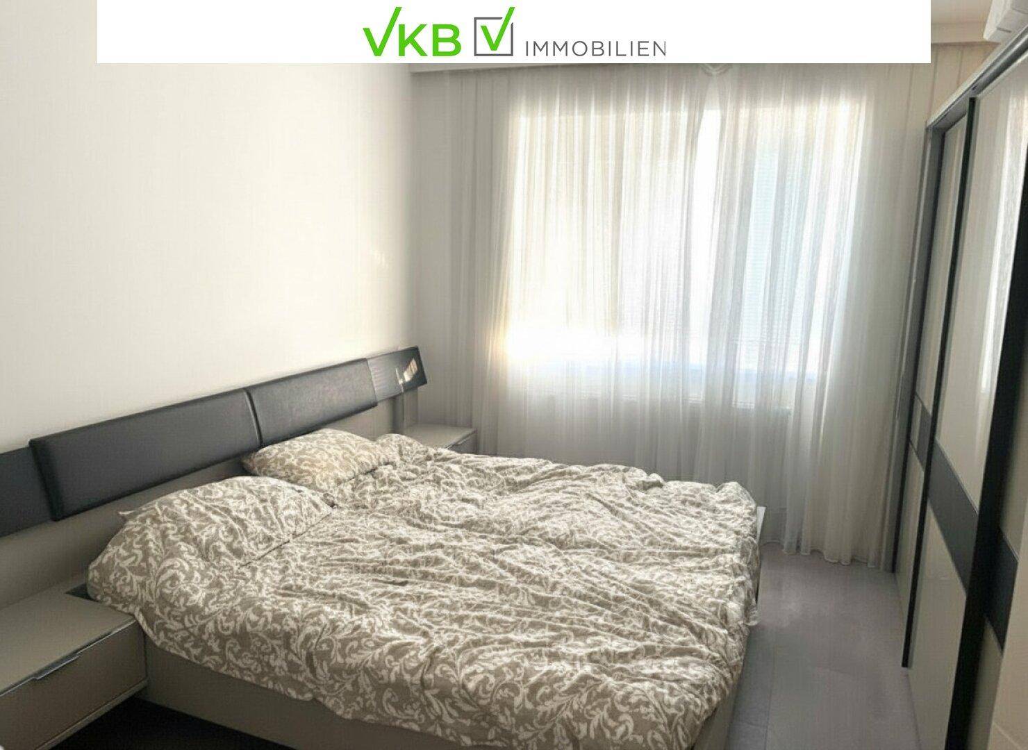 Helles Schlafzimmer mit Doppelbett, Nachttischen und großem Fenster mit Vorhängen.