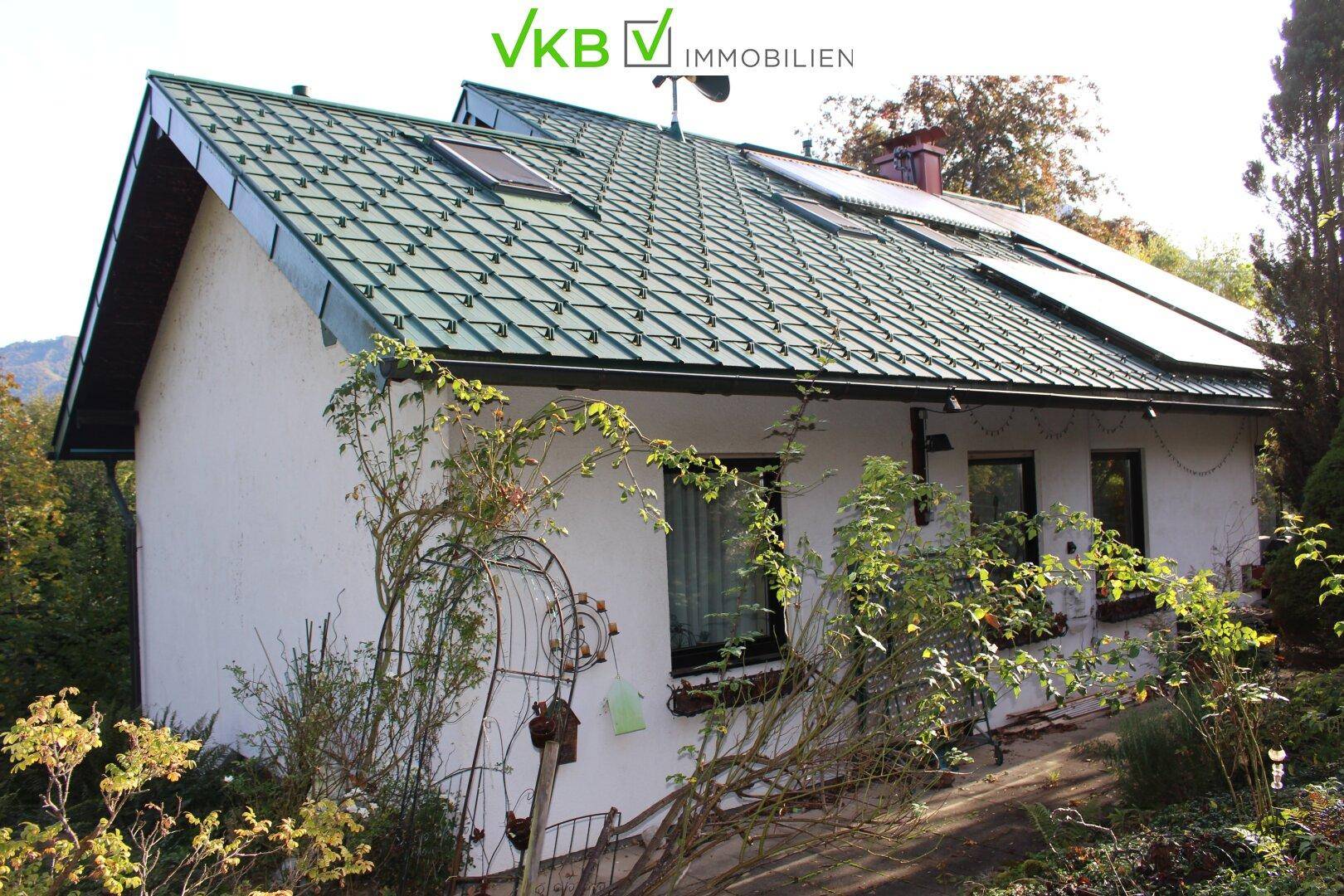Seitenansicht eines weißen Hauses mit grünem Satteldach und Photovoltaikanlage, umgeben von Pflanzen.