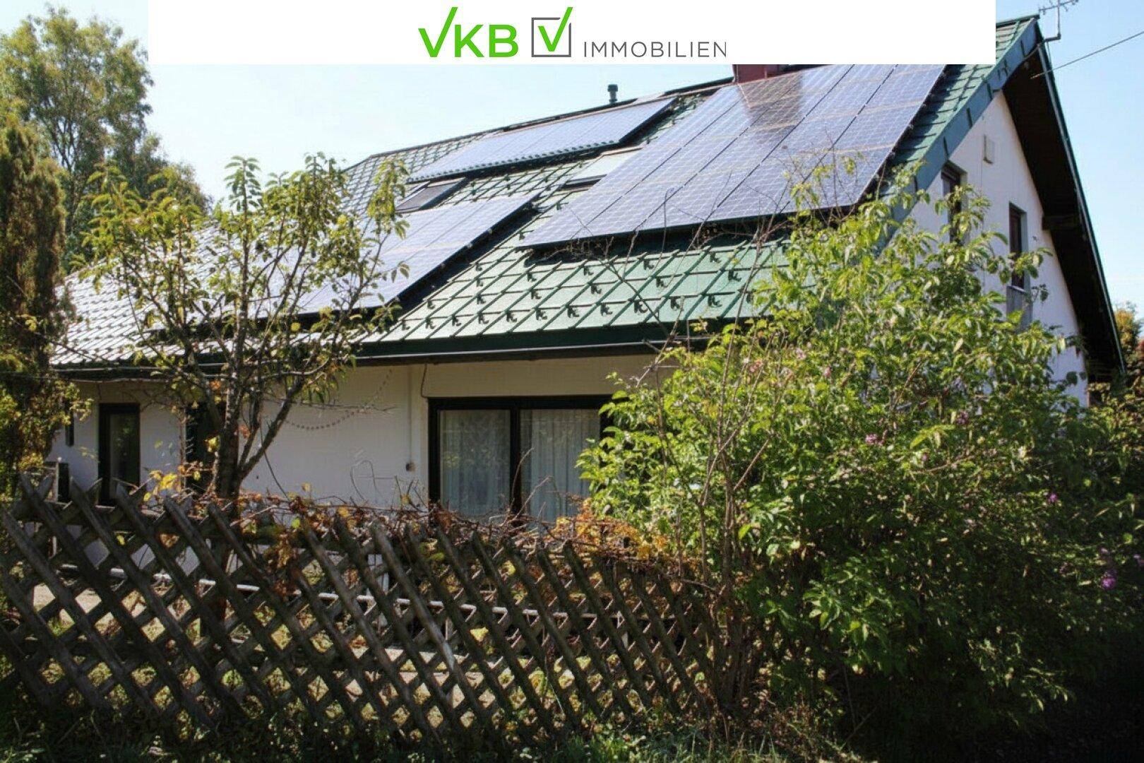 Haus mit grünem Satteldach und Photovoltaikanlage, umgeben von einem Holzzaun und Bäumen.