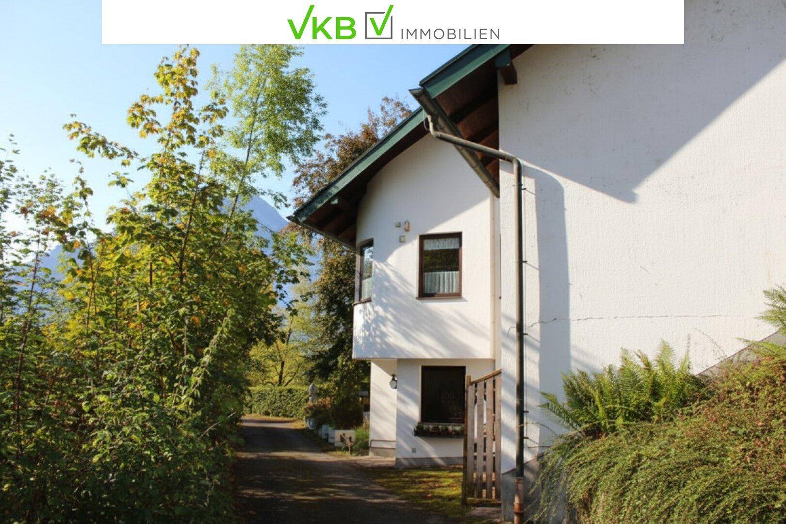 Seitenansicht eines weißen Hauses mit grünem Dach und einer Zufahrt, umgeben von Bäumen.