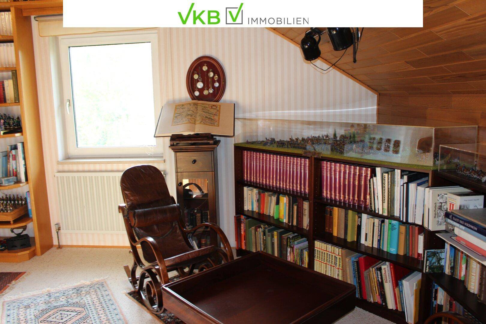 Wohnbereich mit Bücherregalen, Schaukelstuhl und einem Fenster, das Tageslicht hereinlässt.