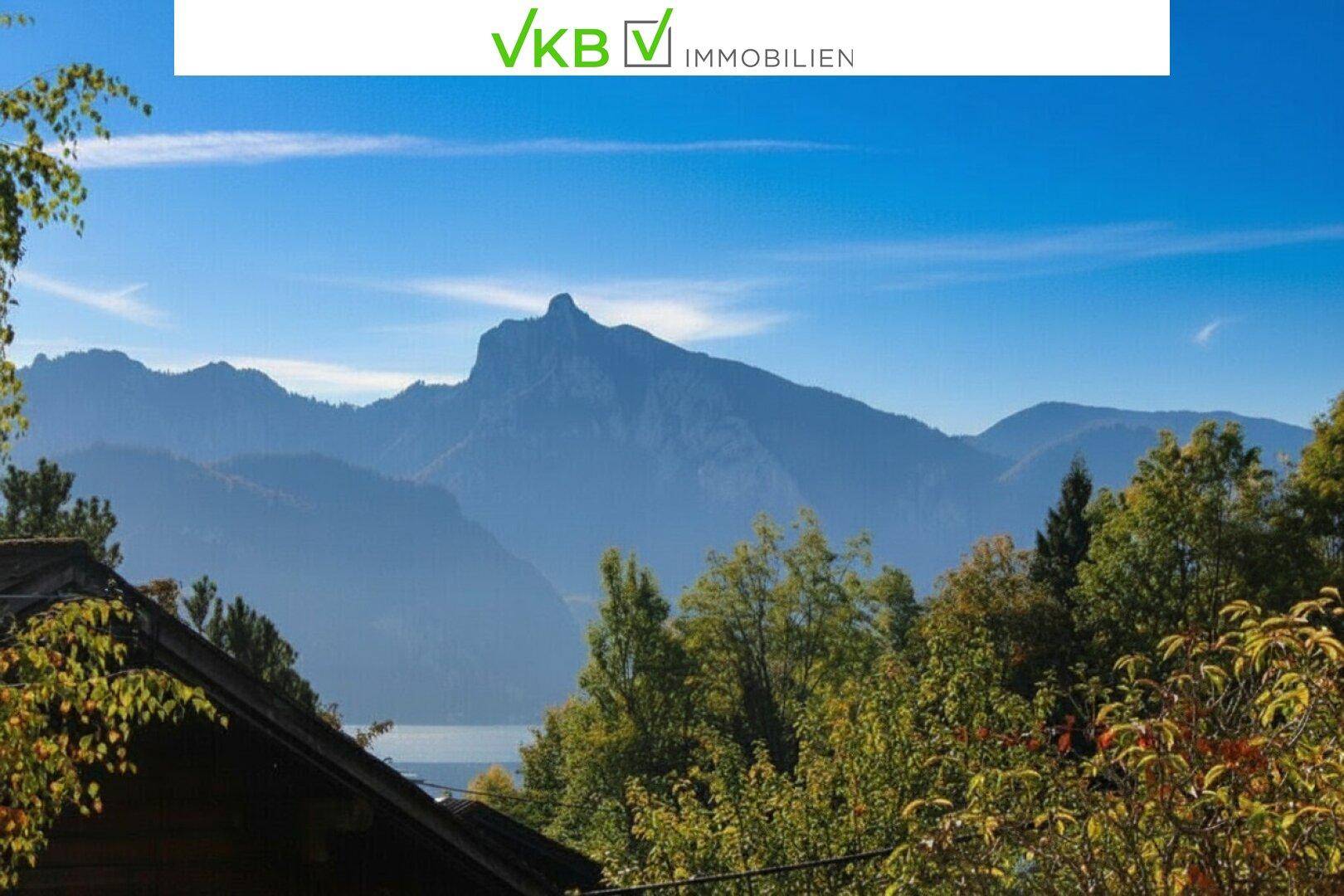 Panoramablick auf majestätische Berge und einen glitzernden See, umgeben von üppiger Natur und blauem Himmel.