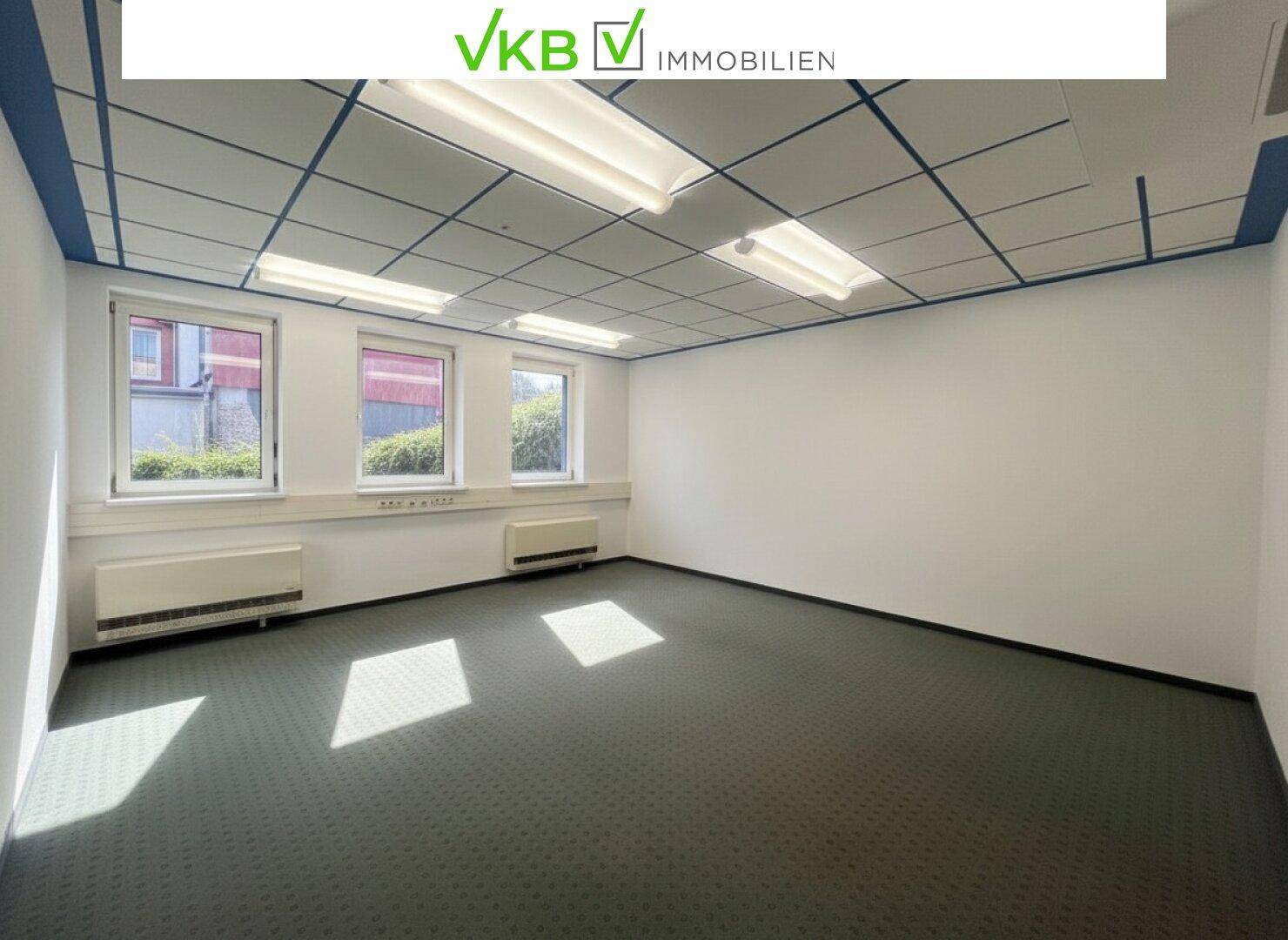 Leerer Büroraum mit Teppichboden, Rasterdecke und drei Fenstern, die Tageslicht hereinlassen.
