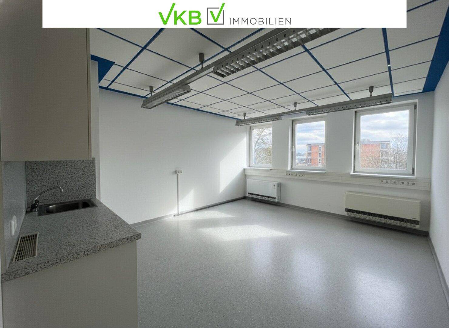 Heller Büroraum mit drei Fenstern, die Tageslicht hereinlassen, und einer kleinen Küchenzeile.