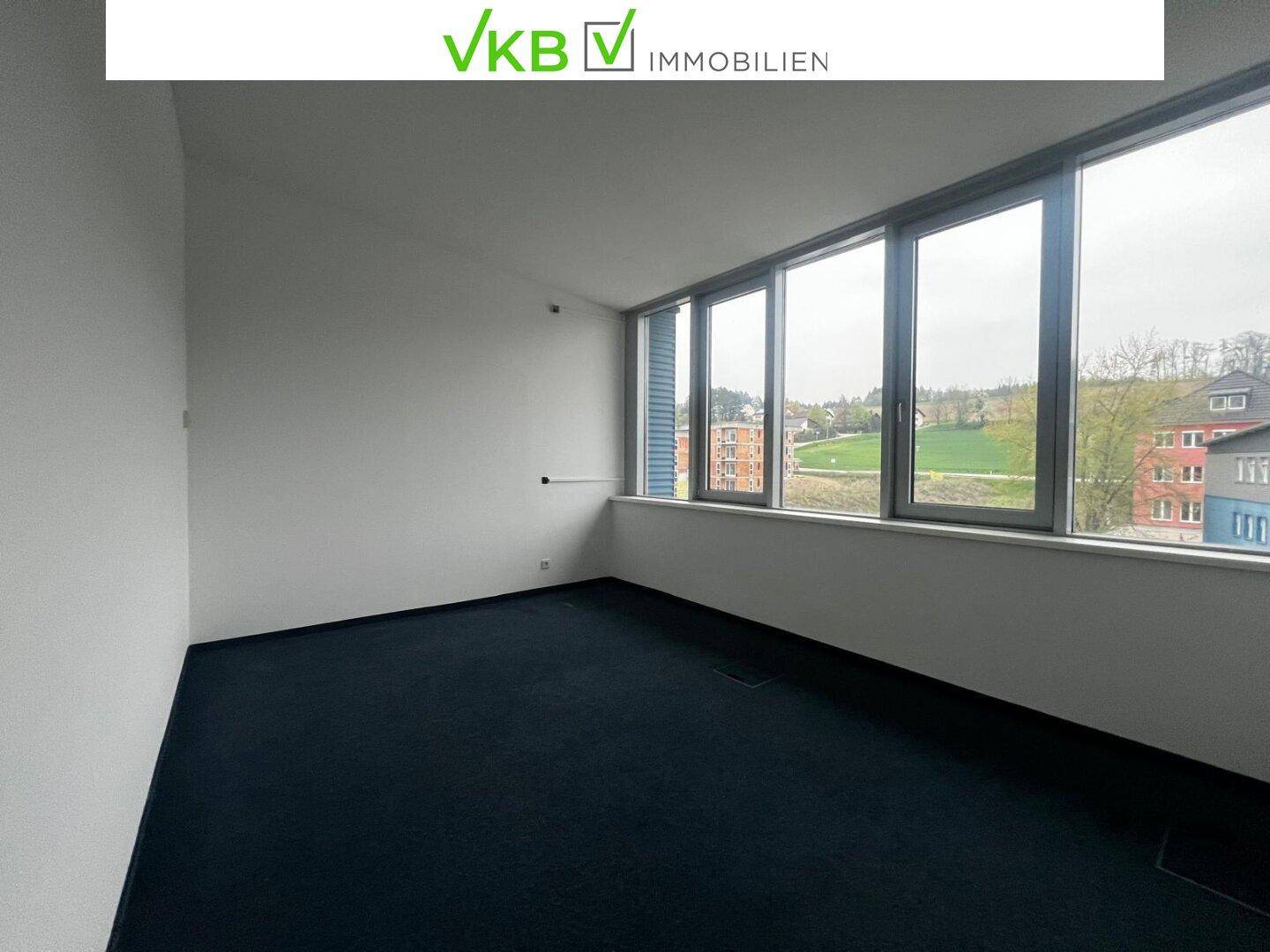 Heller Büroraum mit großen Fenstern, die einen weiten Blick auf die Umgebung bieten, und dunklem Teppichboden.