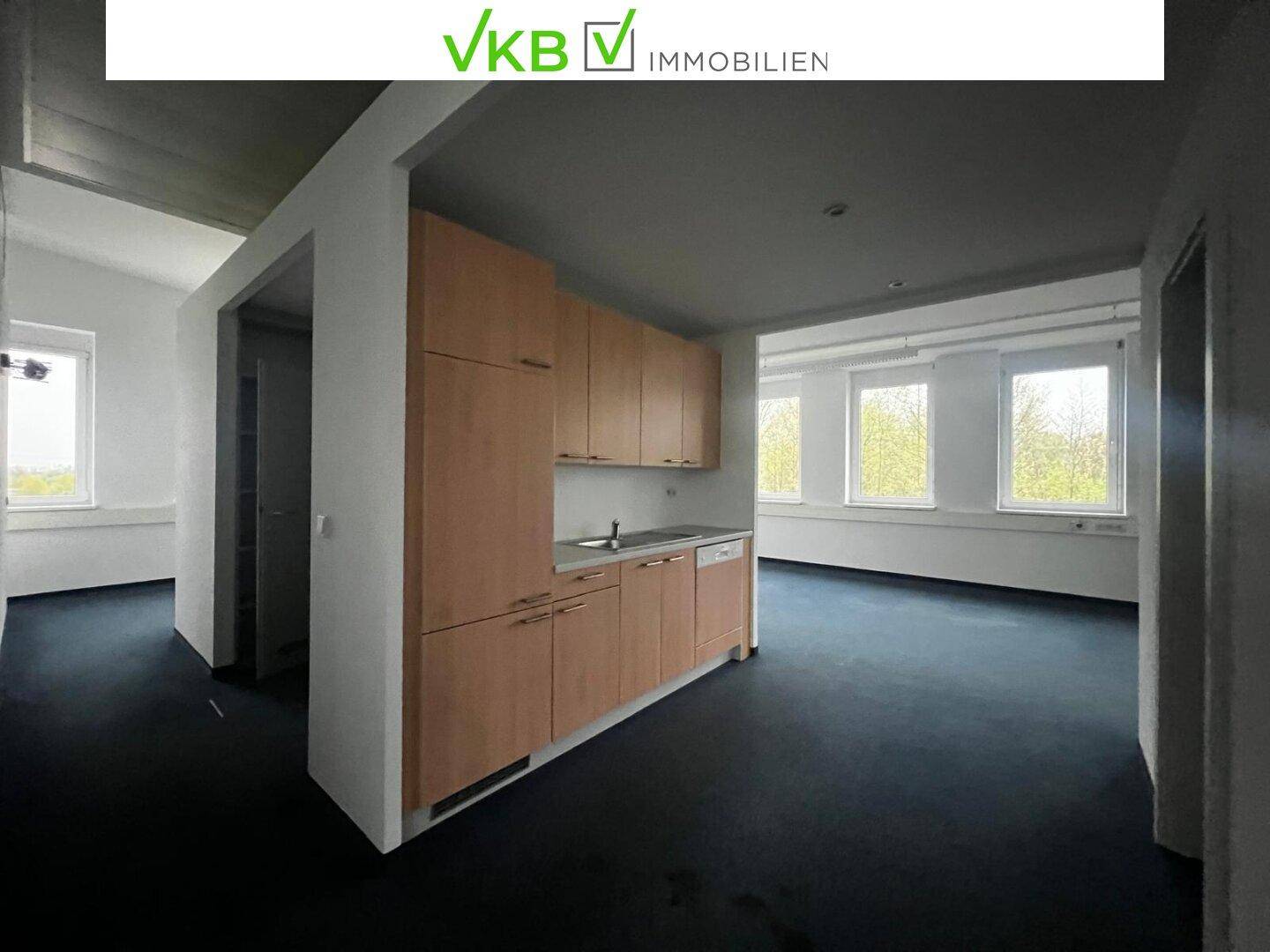 Heller Büroraum mit integrierter Küchenzeile, dunklem Teppichboden und großen Fenstern.