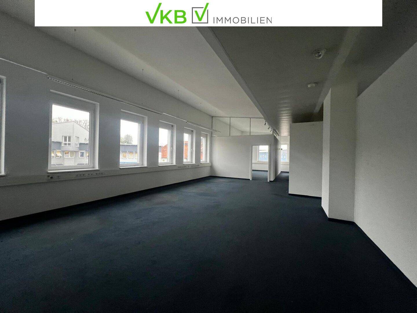Geräumiger, leerer Büroraum mit dunklem Teppichboden und mehreren Fenstern, bereit zur individuellen Gestaltung.