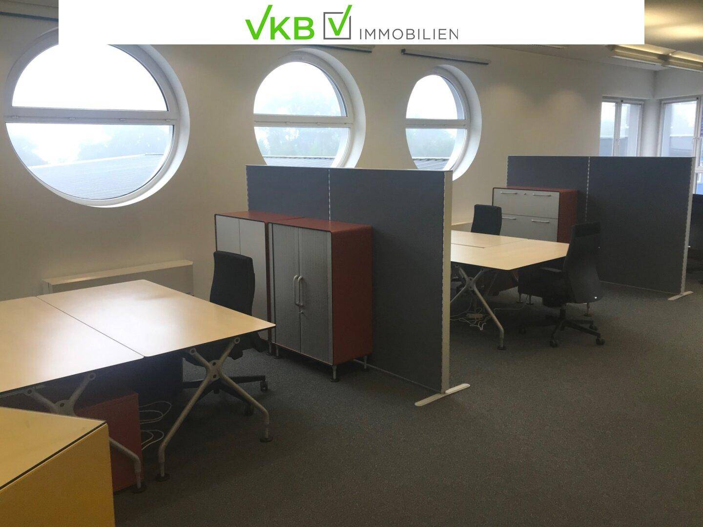 Heller Büroraum mit runden Fenstern, Teppichboden und modernen Arbeitsplatzteilern.