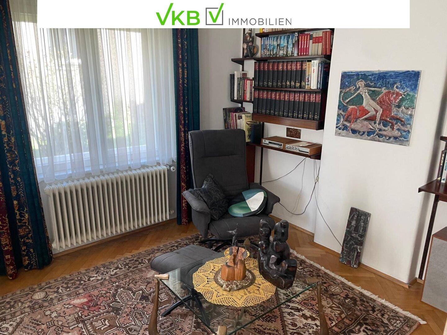 Gemütliches Wohnzimmer mit Sessel, Couchtisch, Bücherregal und großem Fenster.