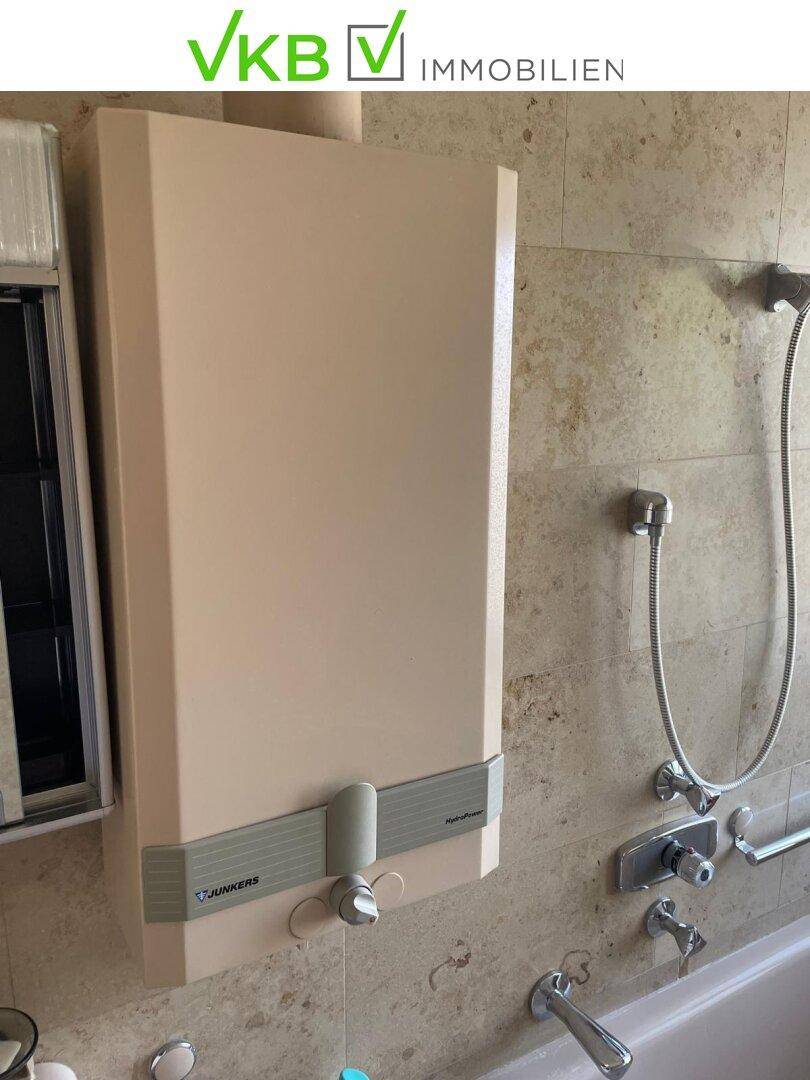 Detailansicht des Badezimmers mit Warmwasserbereiter, Duscharmatur und beigefarbenen Fliesen.