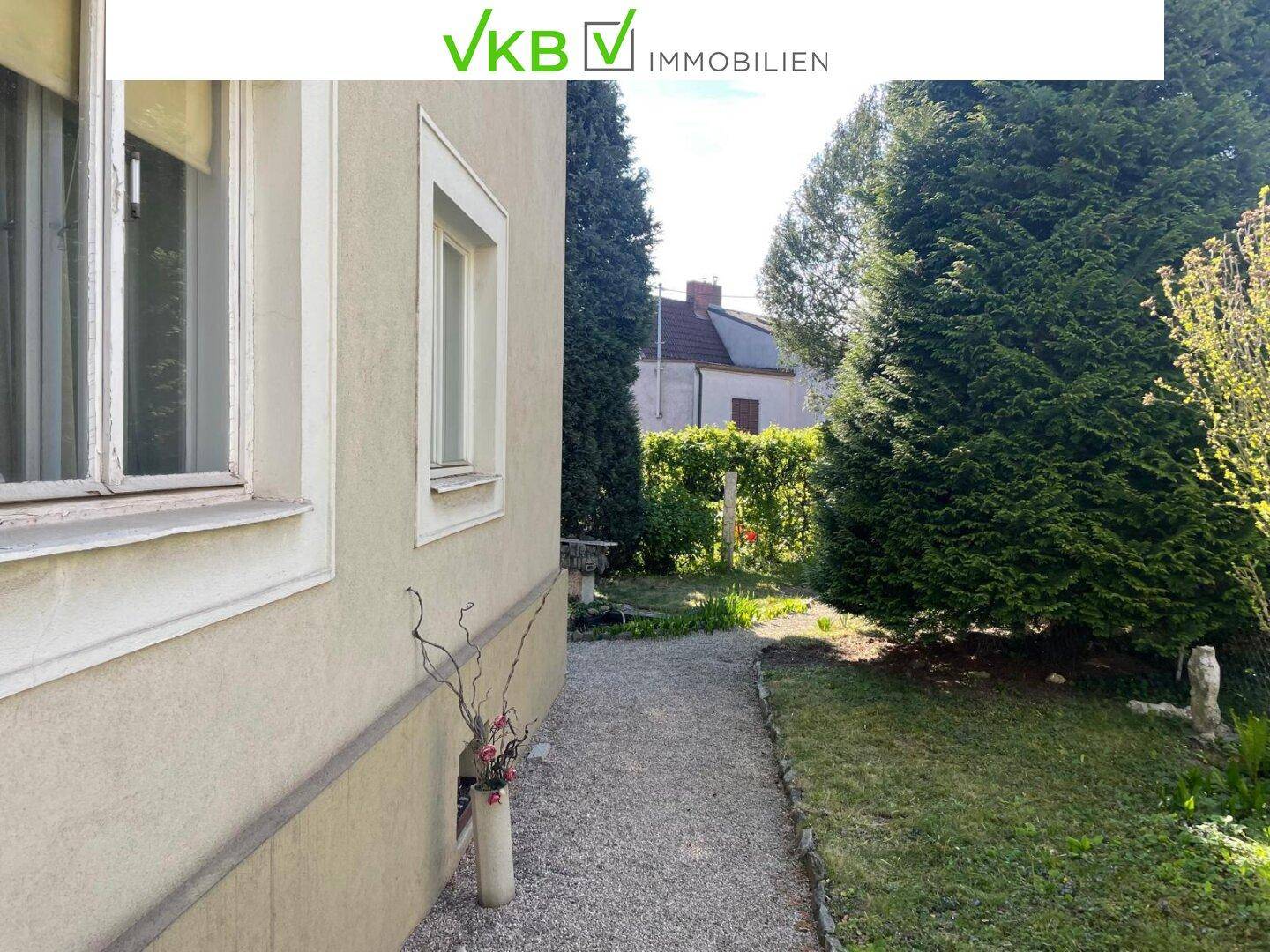 Seitenansicht eines Hauses mit Kiesweg, Rasenfläche und grünen Büschen unter blauem Himmel.