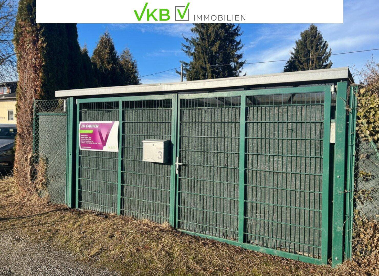 Grüner Metallzaun mit Tor und Briefkasten, der das zum Verkauf stehende Grundstück abgrenzt.