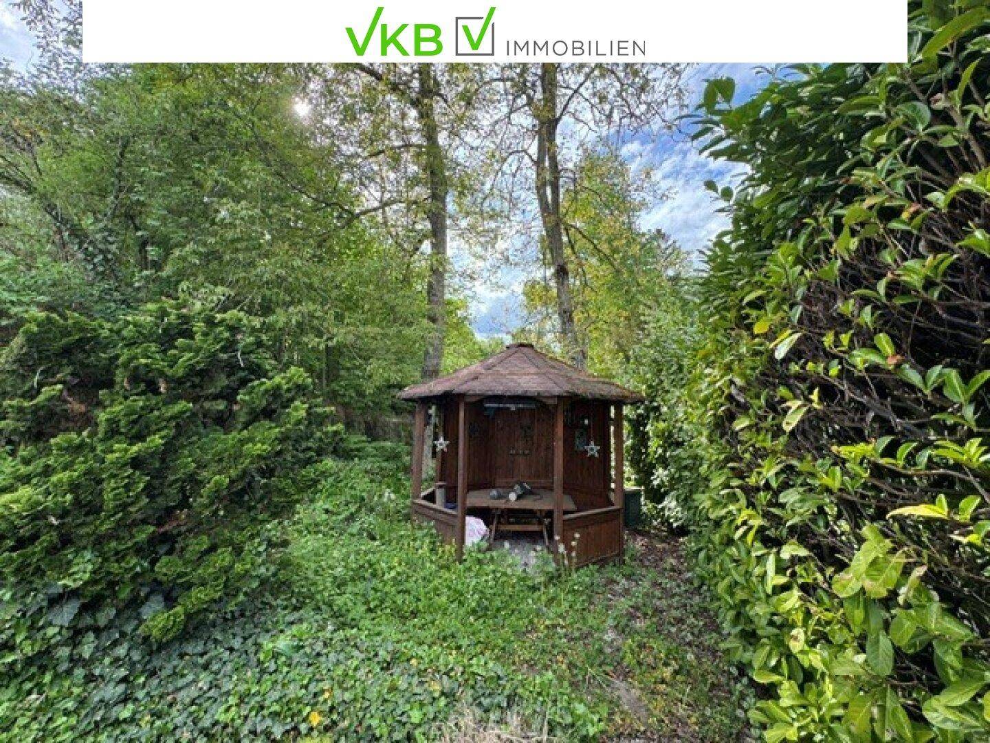 Gemütlicher hölzerner Pavillon im Garten, umgeben von Bäumen und Sträuchern.