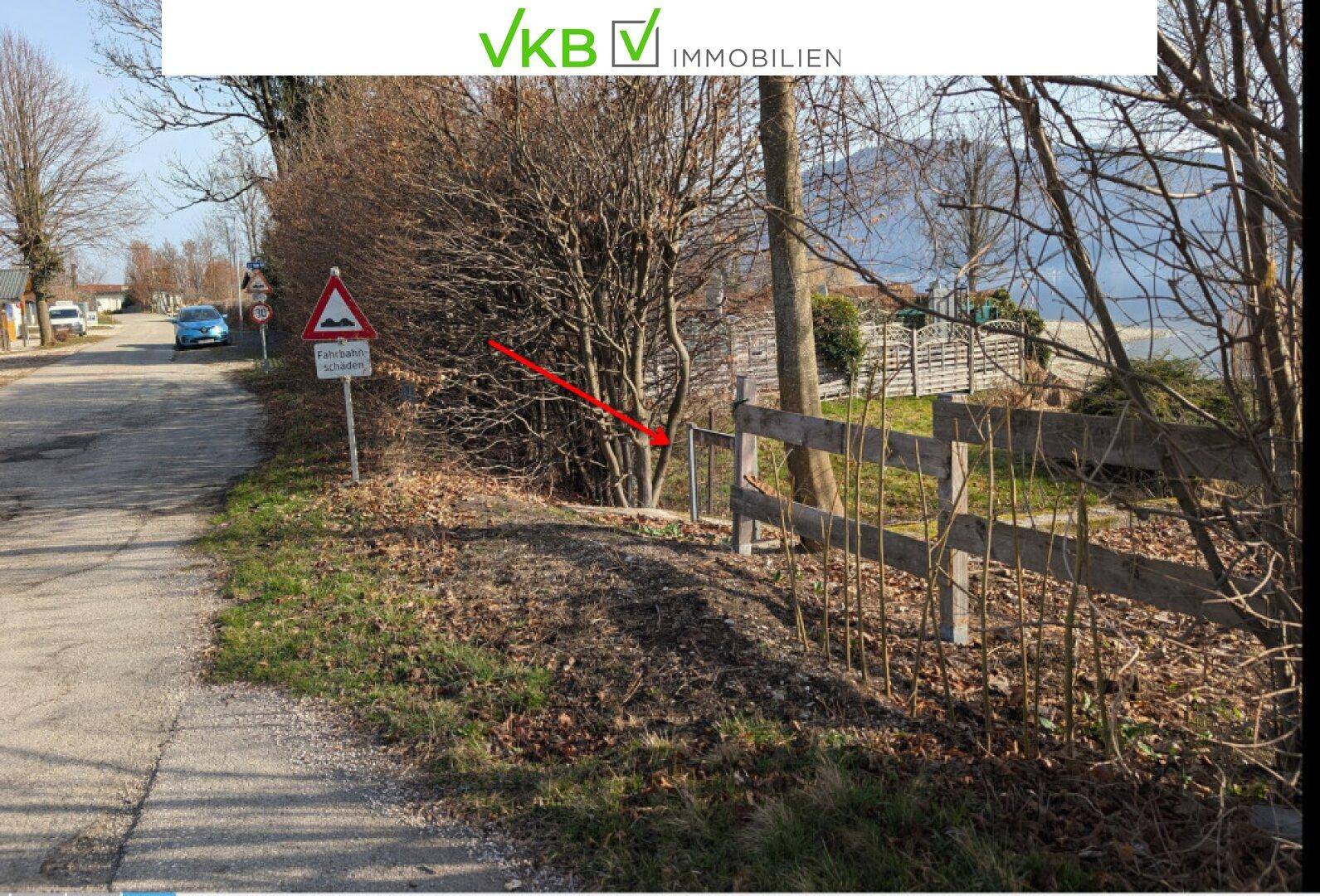 Ländliche Straße mit Holzzaun und Blick auf einen See oder ein Gewässer in der Ferne.