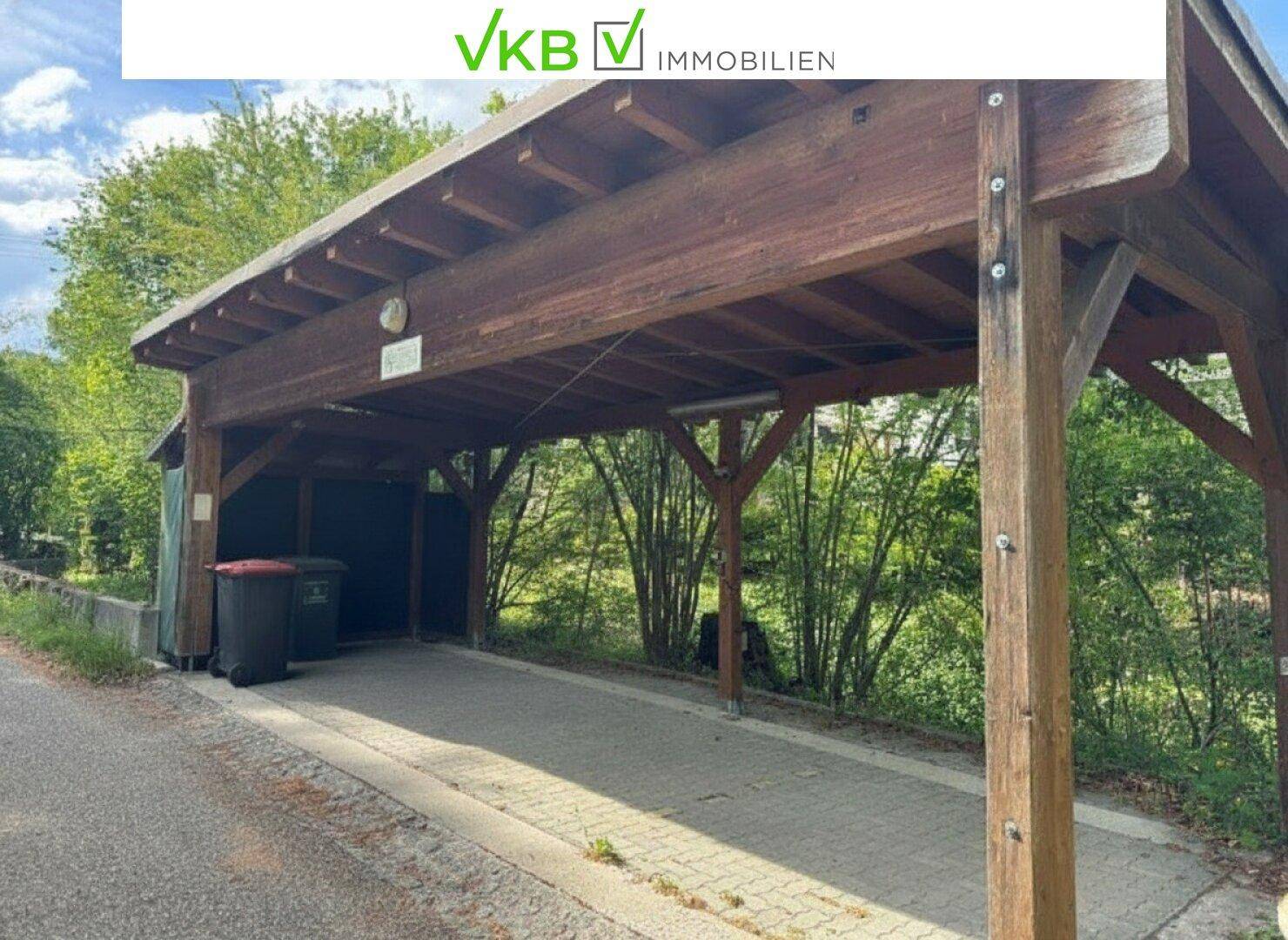 Hölzerner Carport mit Stellplatz für Fahrzeuge und Mülltonnen am Straßenrand.