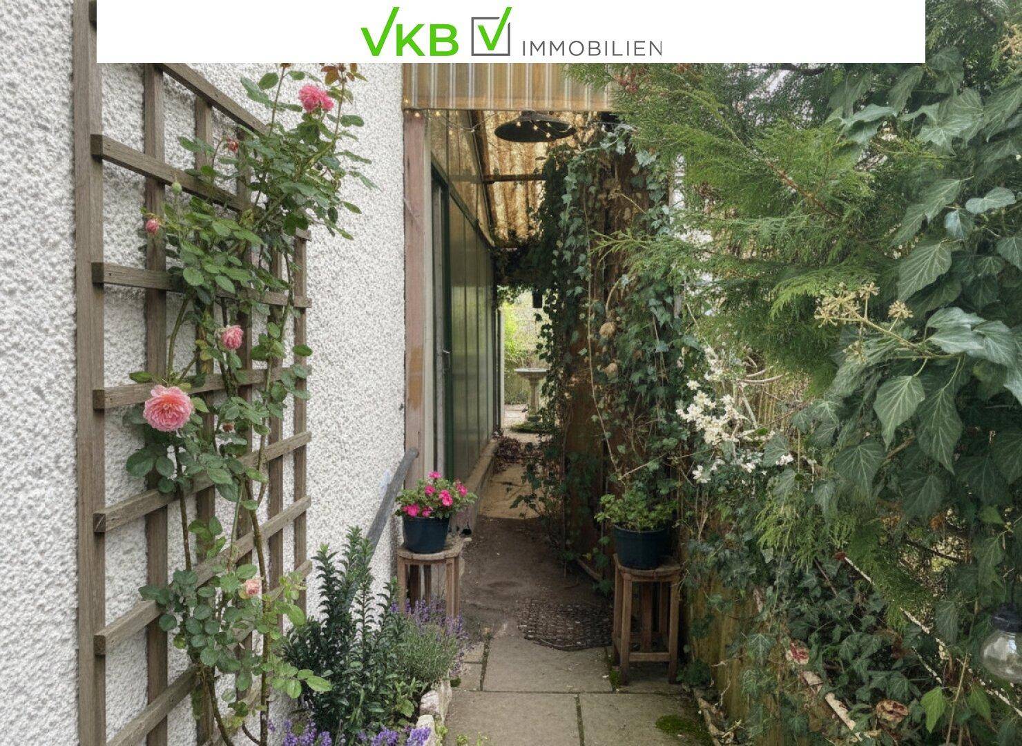 Schmaler Durchgang im Garten mit Kletterrosen und Topfpflanzen entlang der Hauswand.