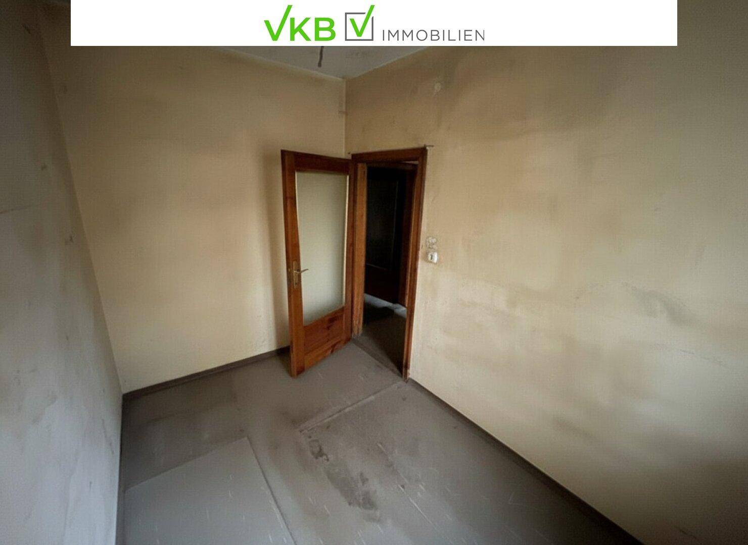 Unmöblierter Raum mit einfacher Holztür und abgenutzten Wänden, sanierungsbedürftig.