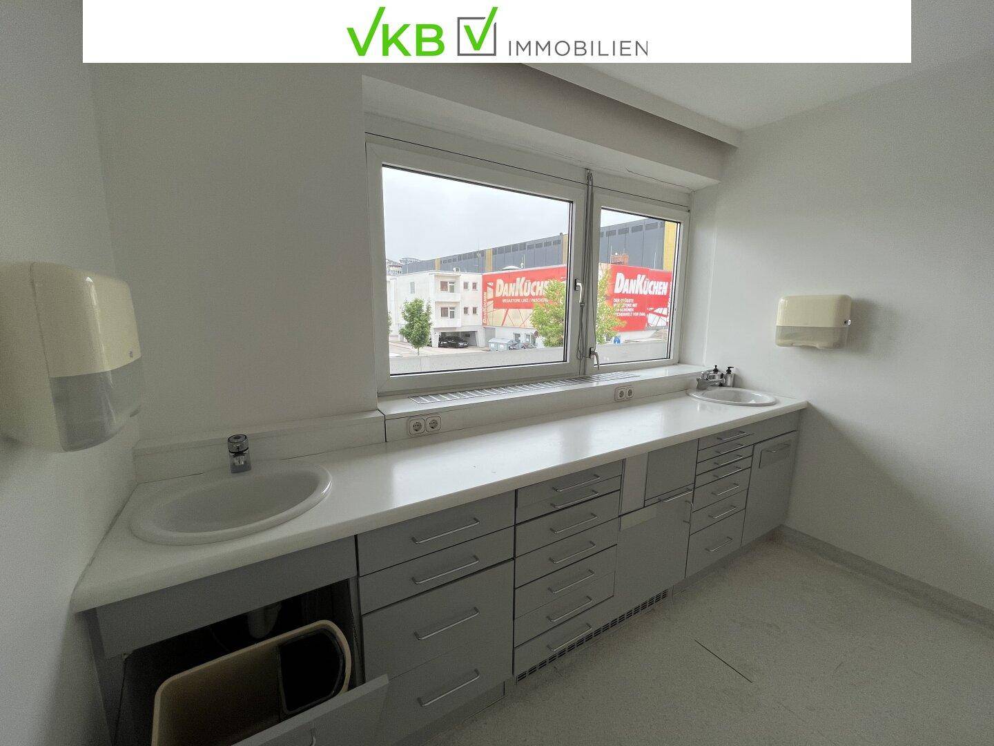 Heller Büroraum mit zwei Waschbecken, langer Arbeitsplatte und großem Fenster mit Stadtblick.