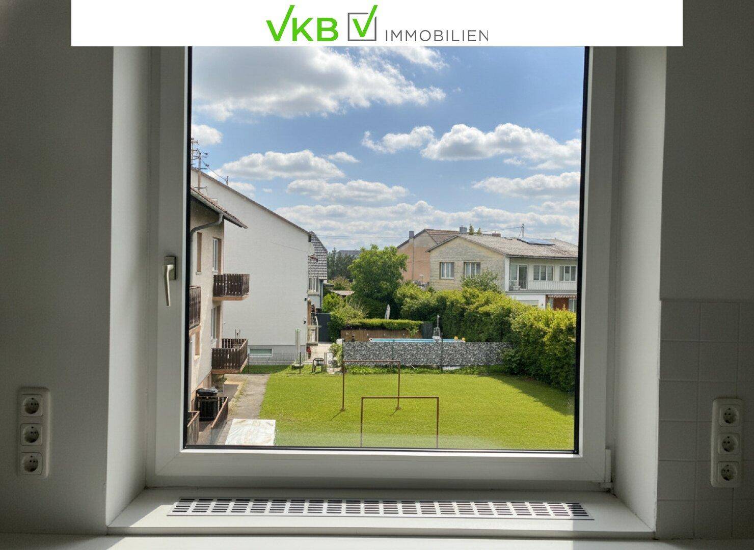 Fenster mit Blick auf einen grünen Garten und benachbarte Häuser unter blauem Himmel.